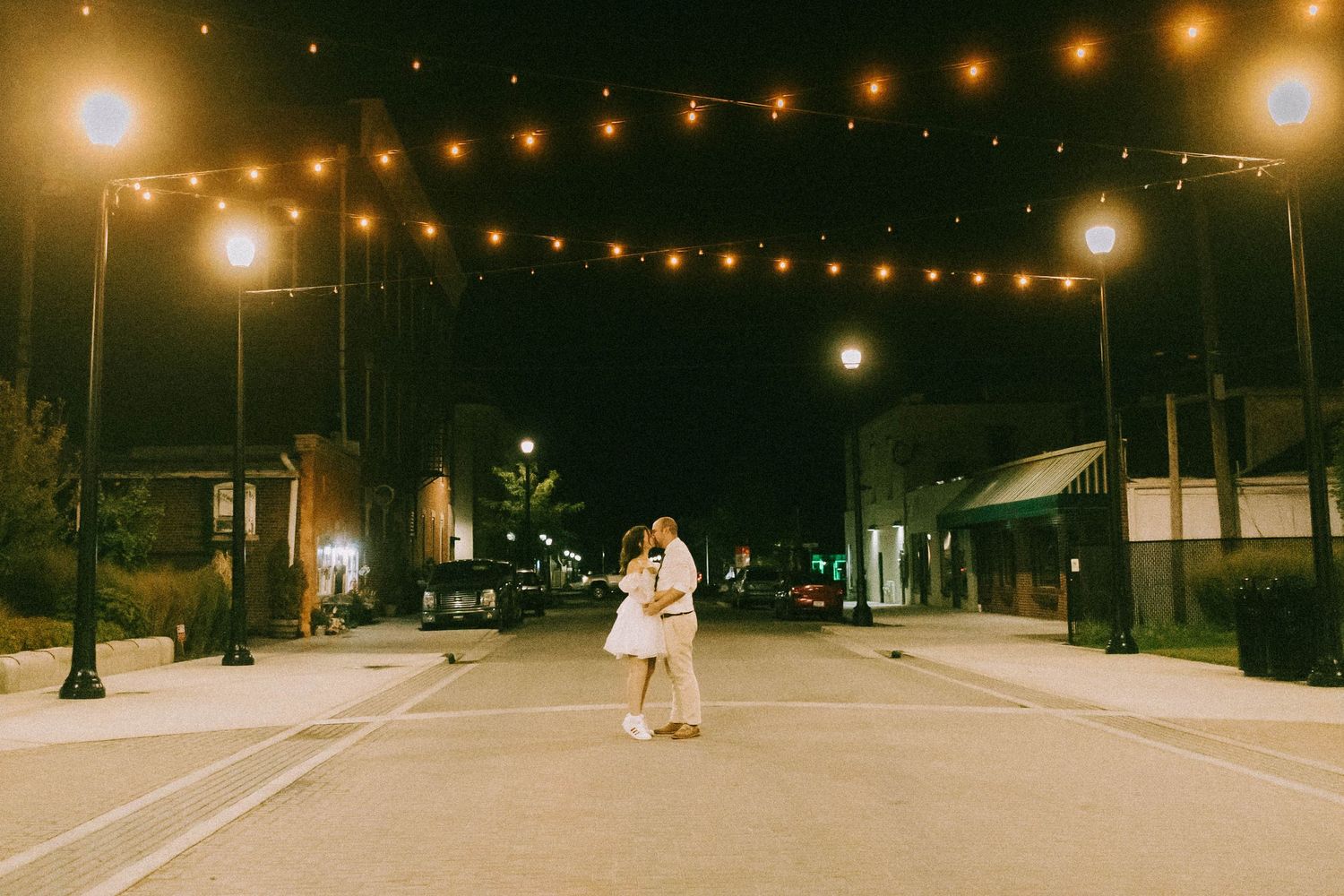 Couple sharing a romantic kiss on an empty street illuminated by string lights at night.