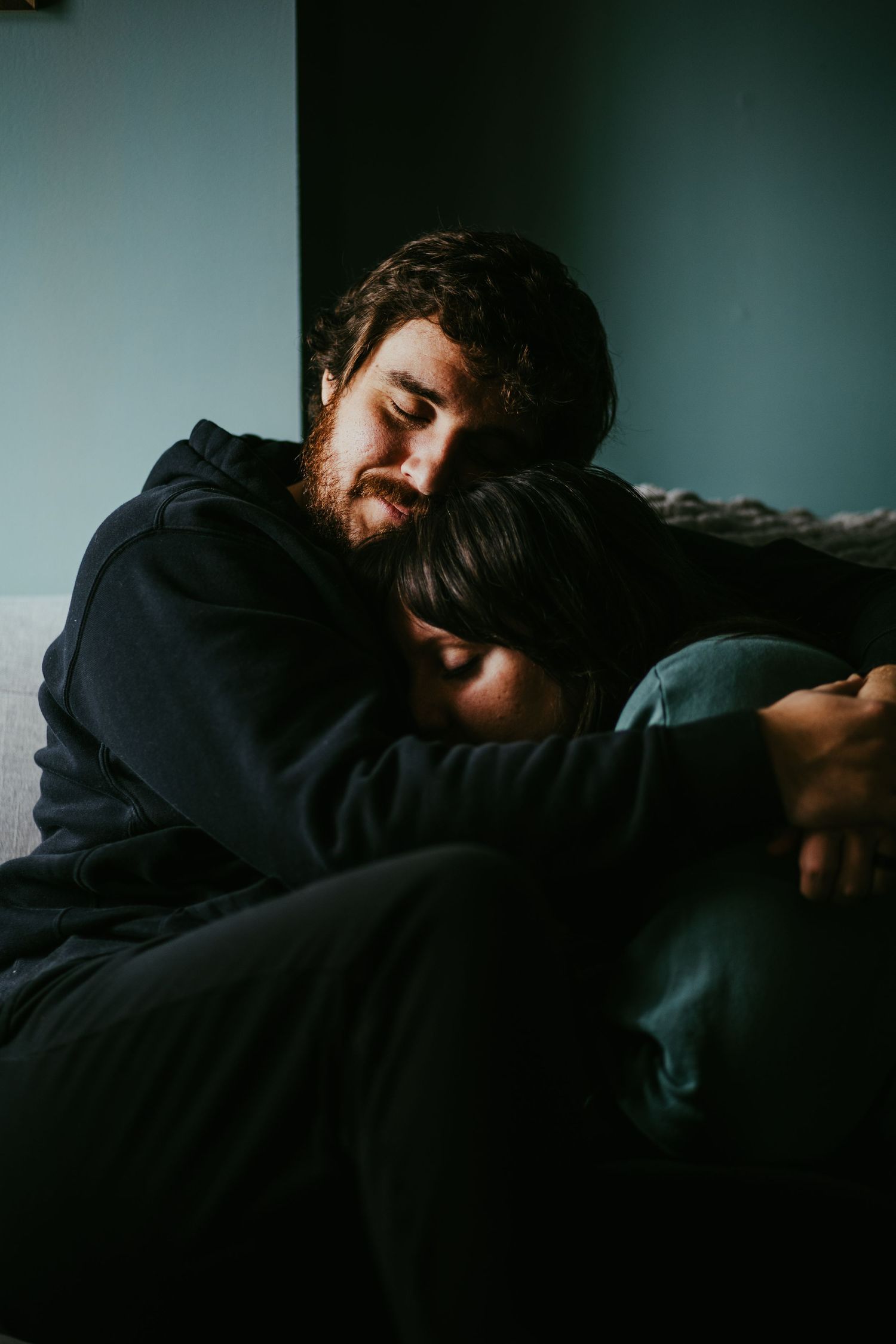 Bearded man tenderly embracing a woman sitting together in a dark moody room.