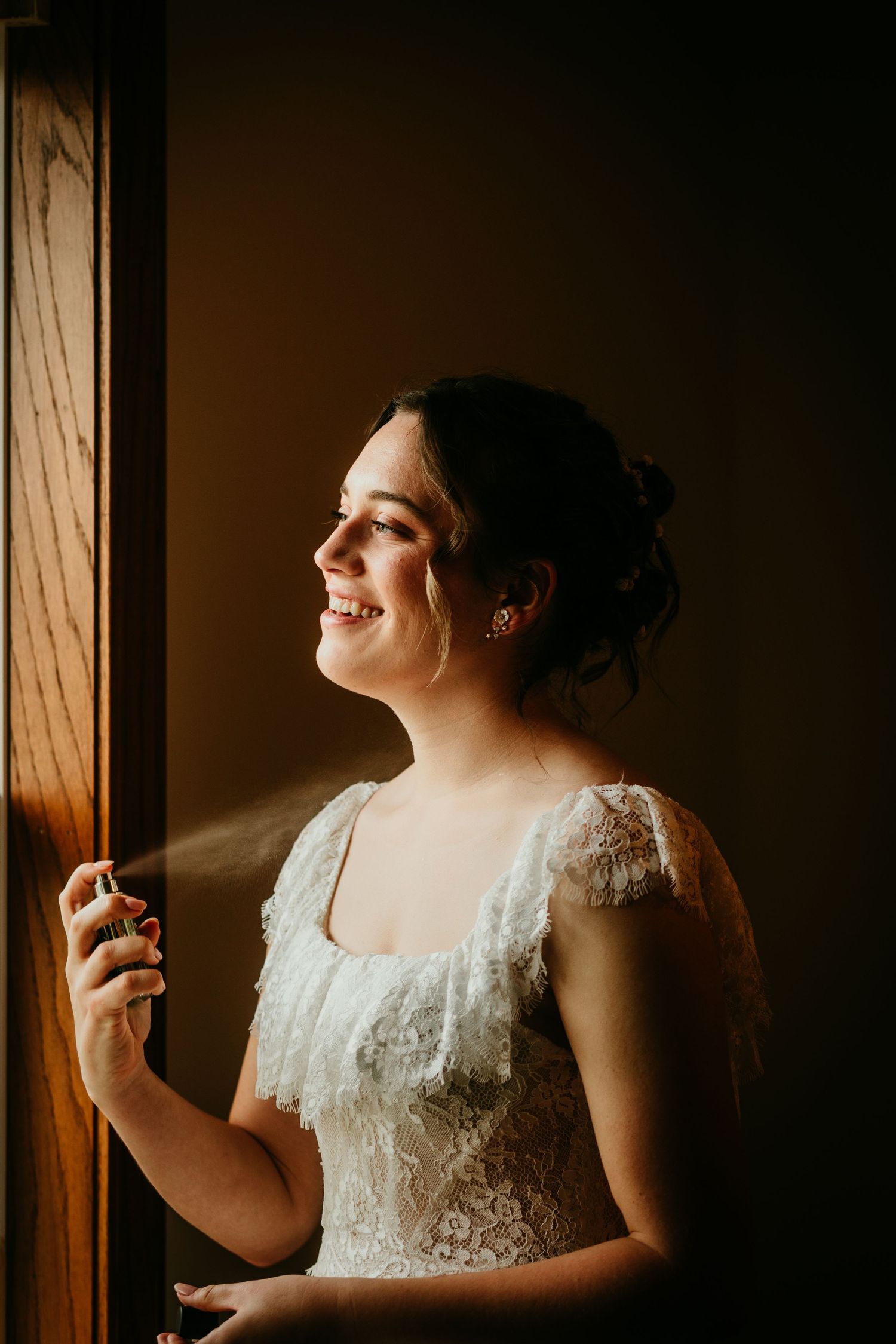 Radiant bride in lace wedding gown spritzing perfume while smiling in warm window light.