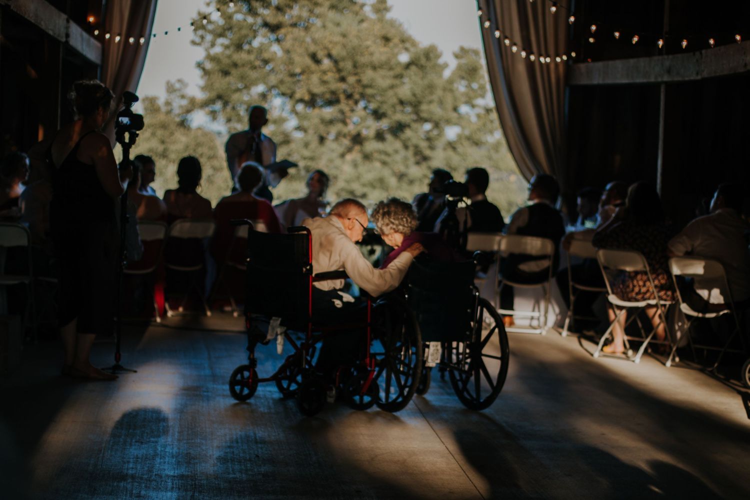 Two people in wheelchairs share a tender moment on a dance floor at an outdoor wedding reception.