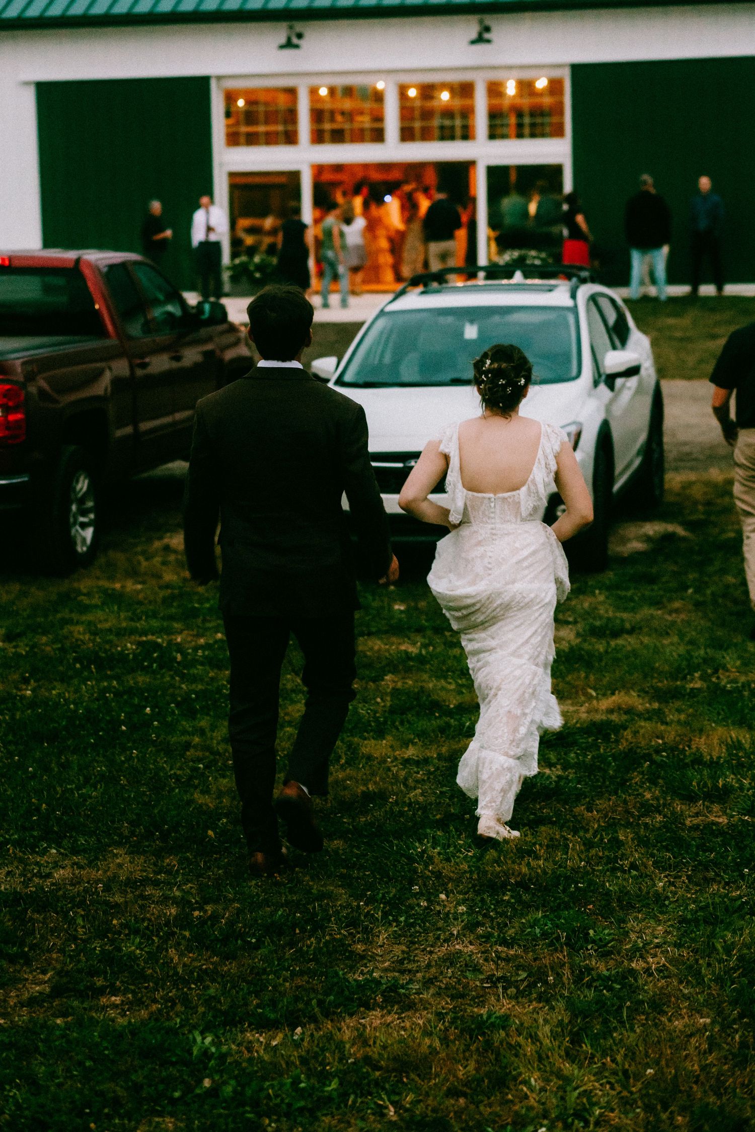 Bride and groom walking hand in hand toward a green barn venue glowing with warm lights.
