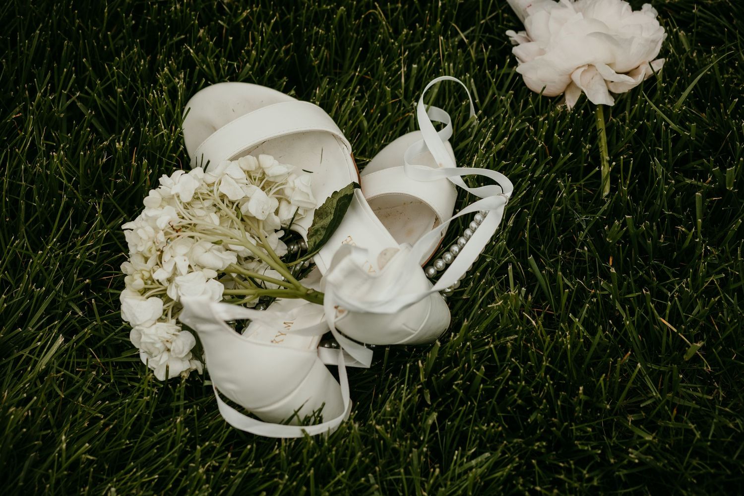 White bridal heels and delicate floral accessories resting on lush green grass before the wedding ceremony.