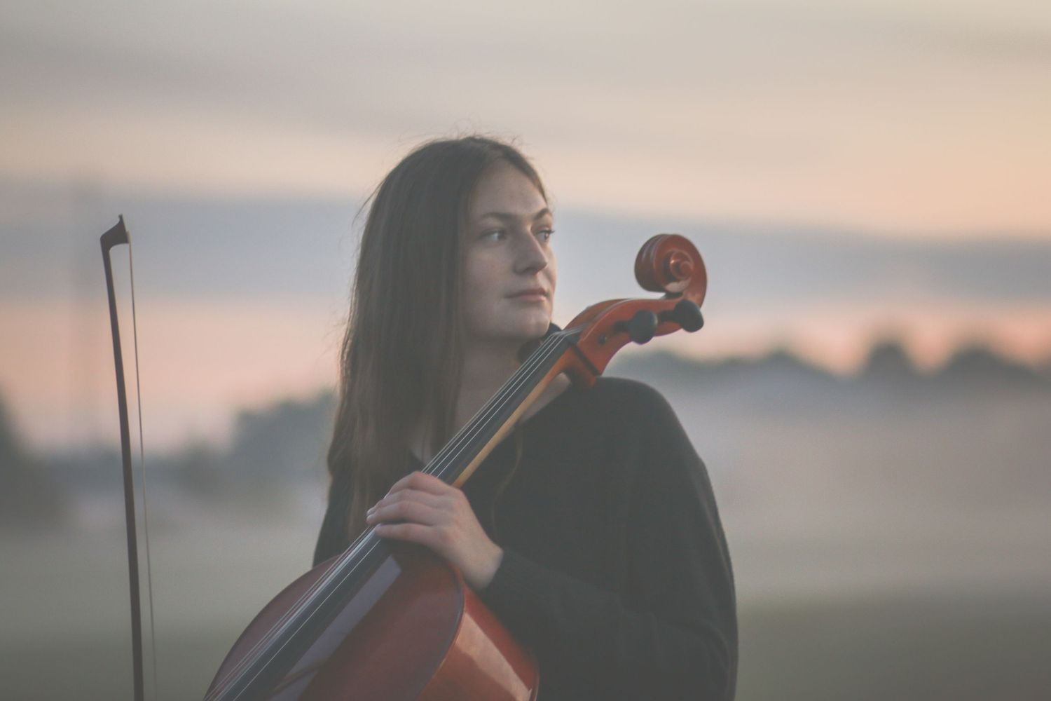 Young woman holding a cello outdoors at dawn with a soft pastel sky behind her.