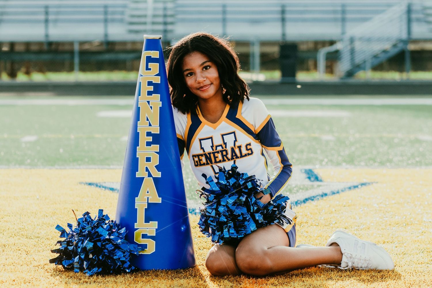 Generals cheerleader posing with blue pom-poms and megaphone on football field in blue and gold uniform.