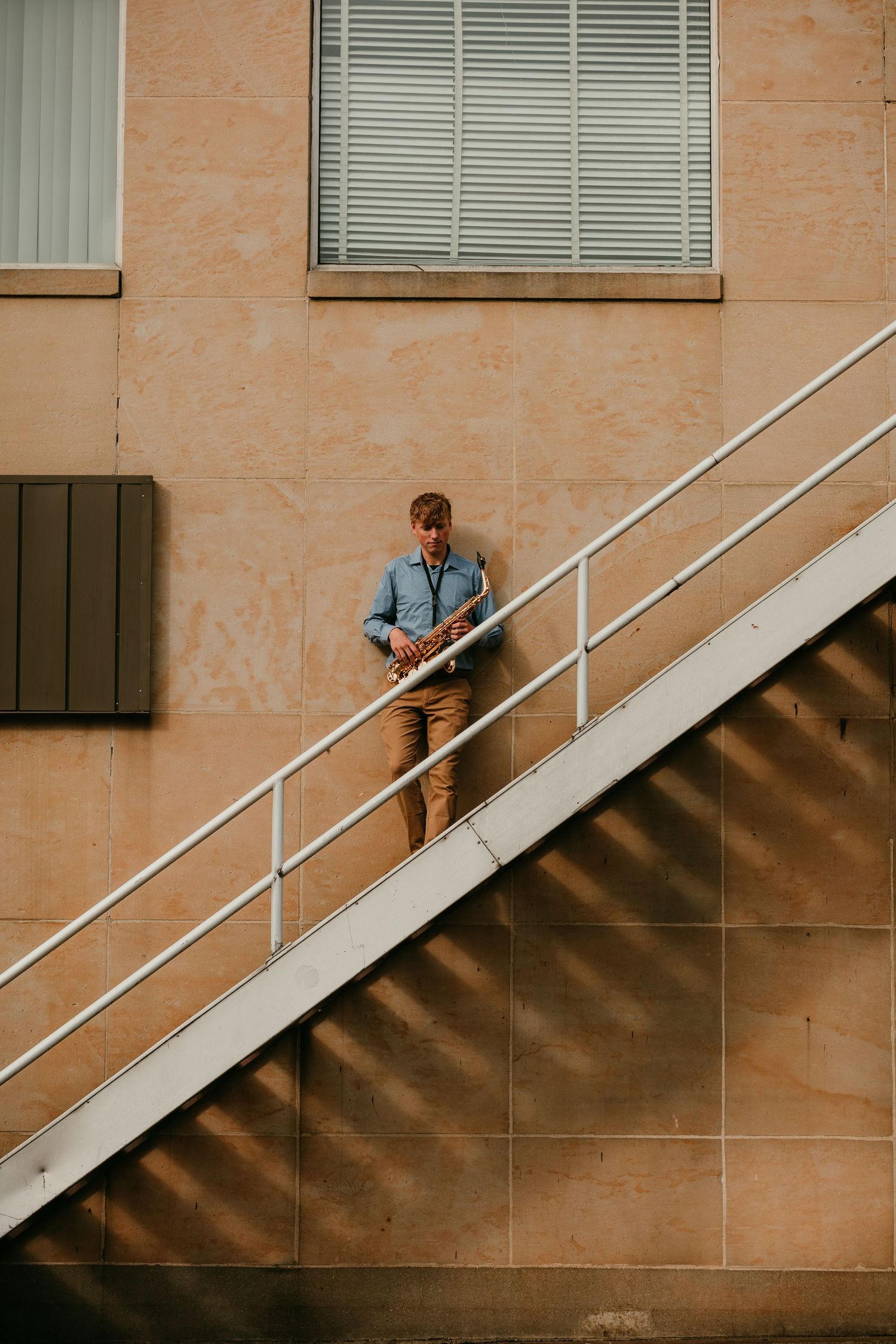Young man playing saxophone on sunlit outdoor staircase against a tan stone building.