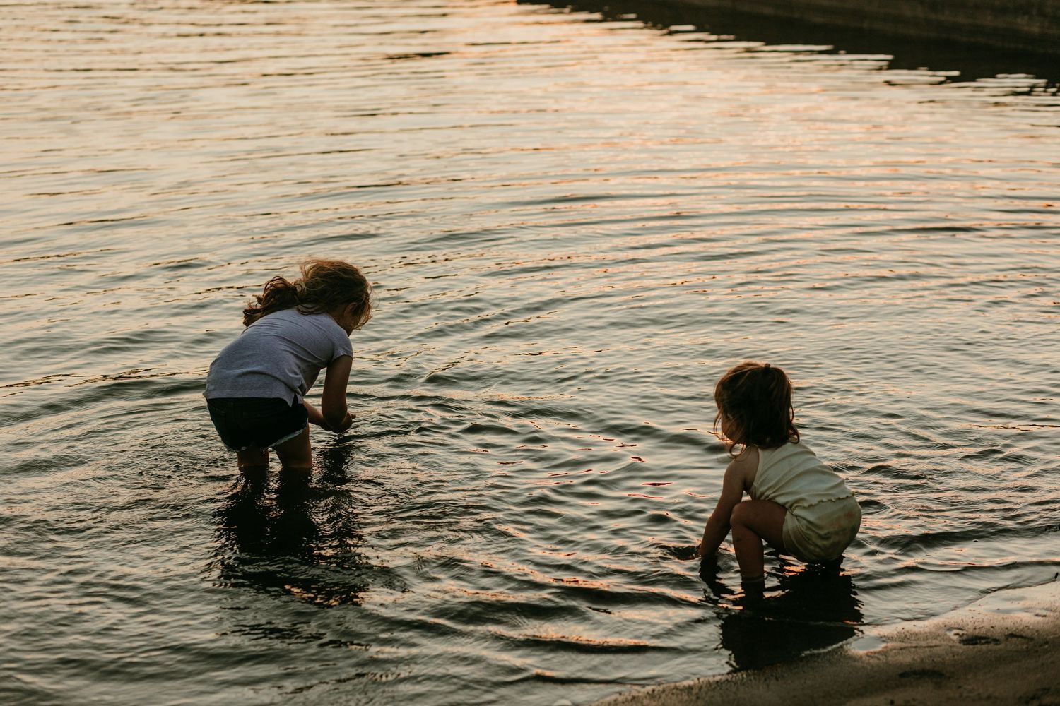 Two young children wading in shallow water at the edge of a lake during golden hour.