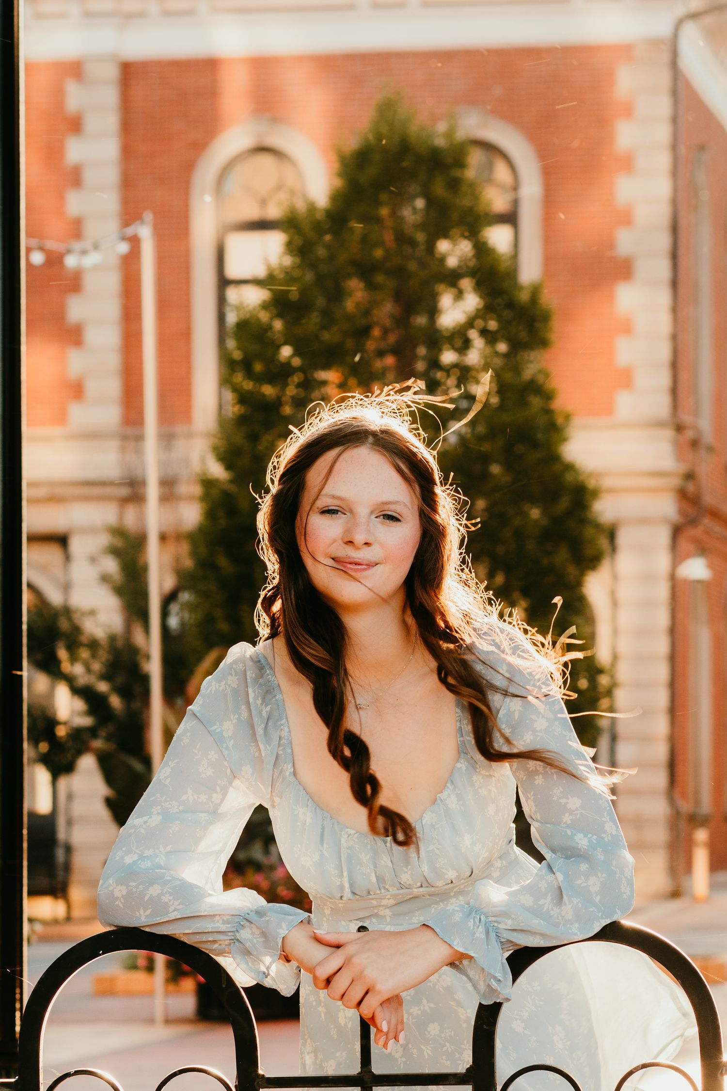 Young woman in a flowy light blue dress leaning on a railing with golden backlight.