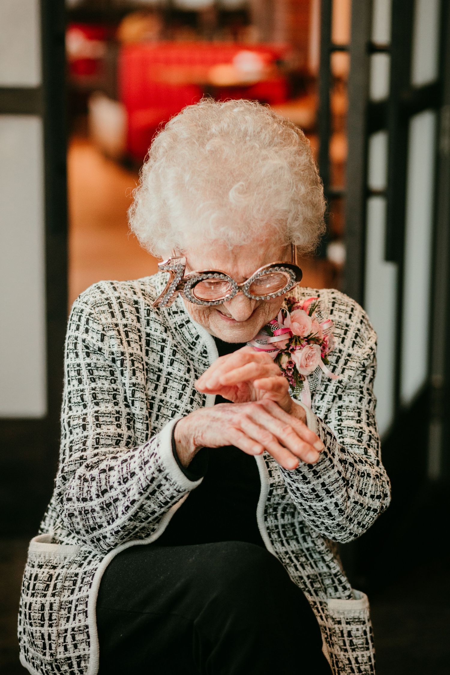 Centenarian woman in tweed jacket examining her floral corsage while wearing decorative novelty glasses.