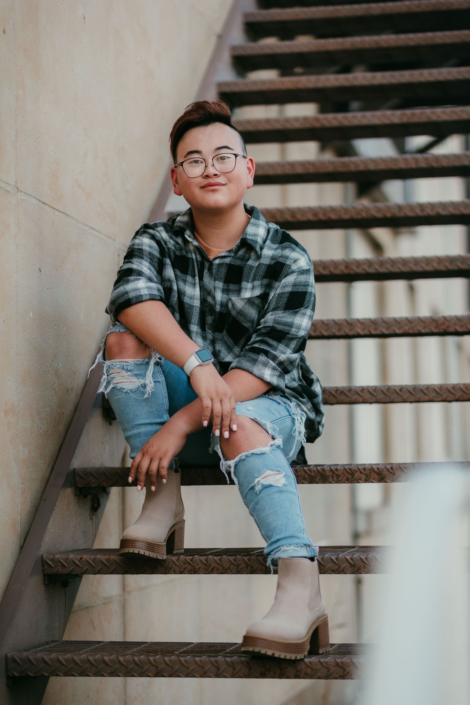 Person in plaid shirt, ripped jeans, and boots sitting on metal stairs, posing confidently for a portrait photo.