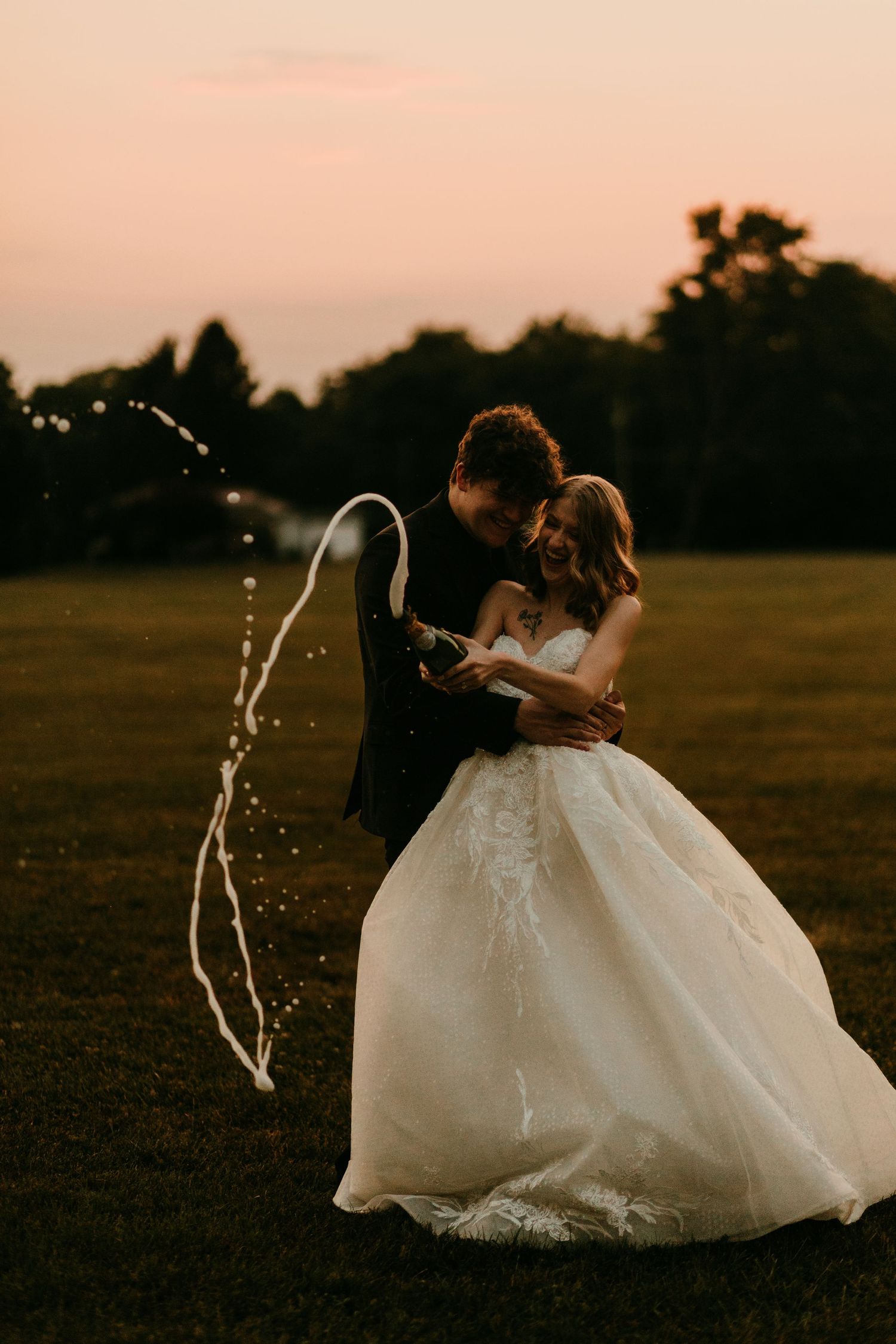 Joyful bride and groom popping champagne at sunset in a lush green field.