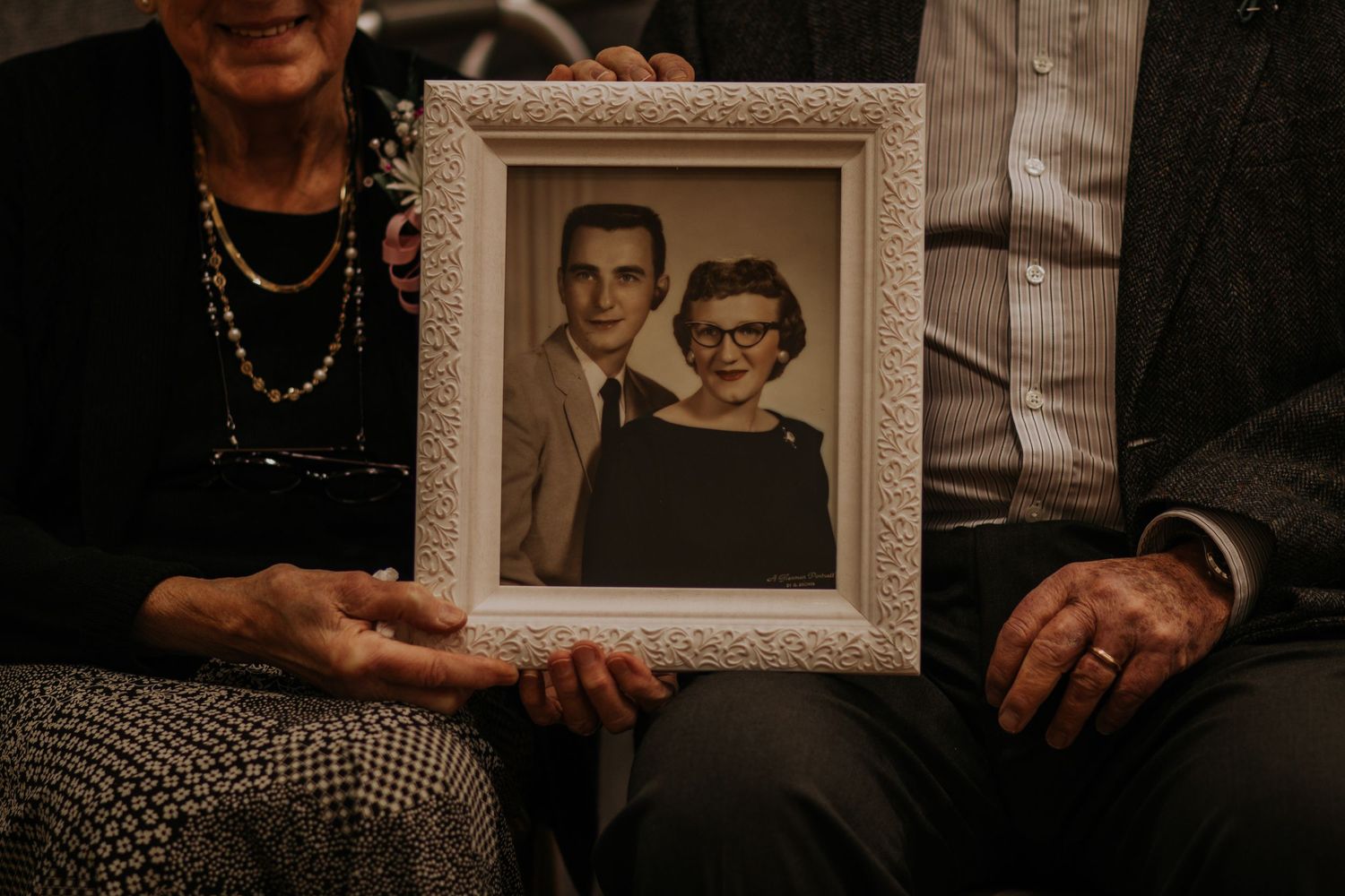 Elderly couple holding a vintage black and white portrait photo in a white decorative frame.
