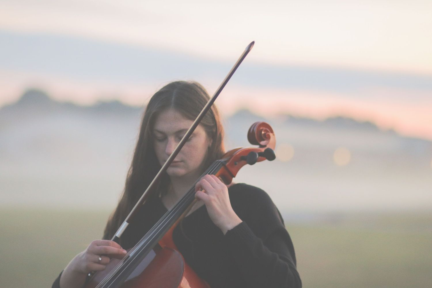 A young woman with long dark hair plays violin outdoors in a misty field at sunset.