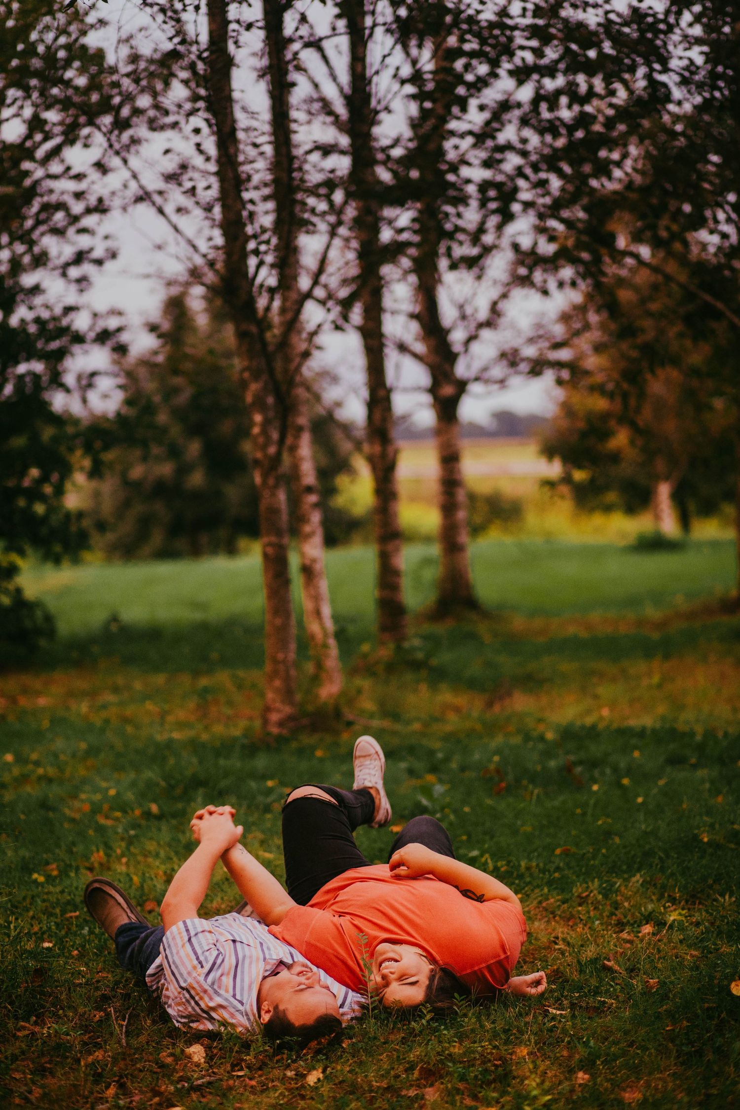 Two children laughing and lying on green grass in an autumn park surrounded by trees.