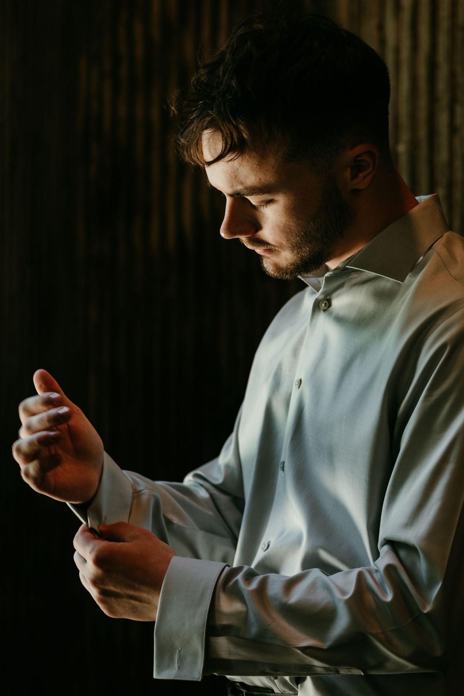 Groom buttoning his shirt cuff in dramatic moody lighting while getting ready for his wedding day.