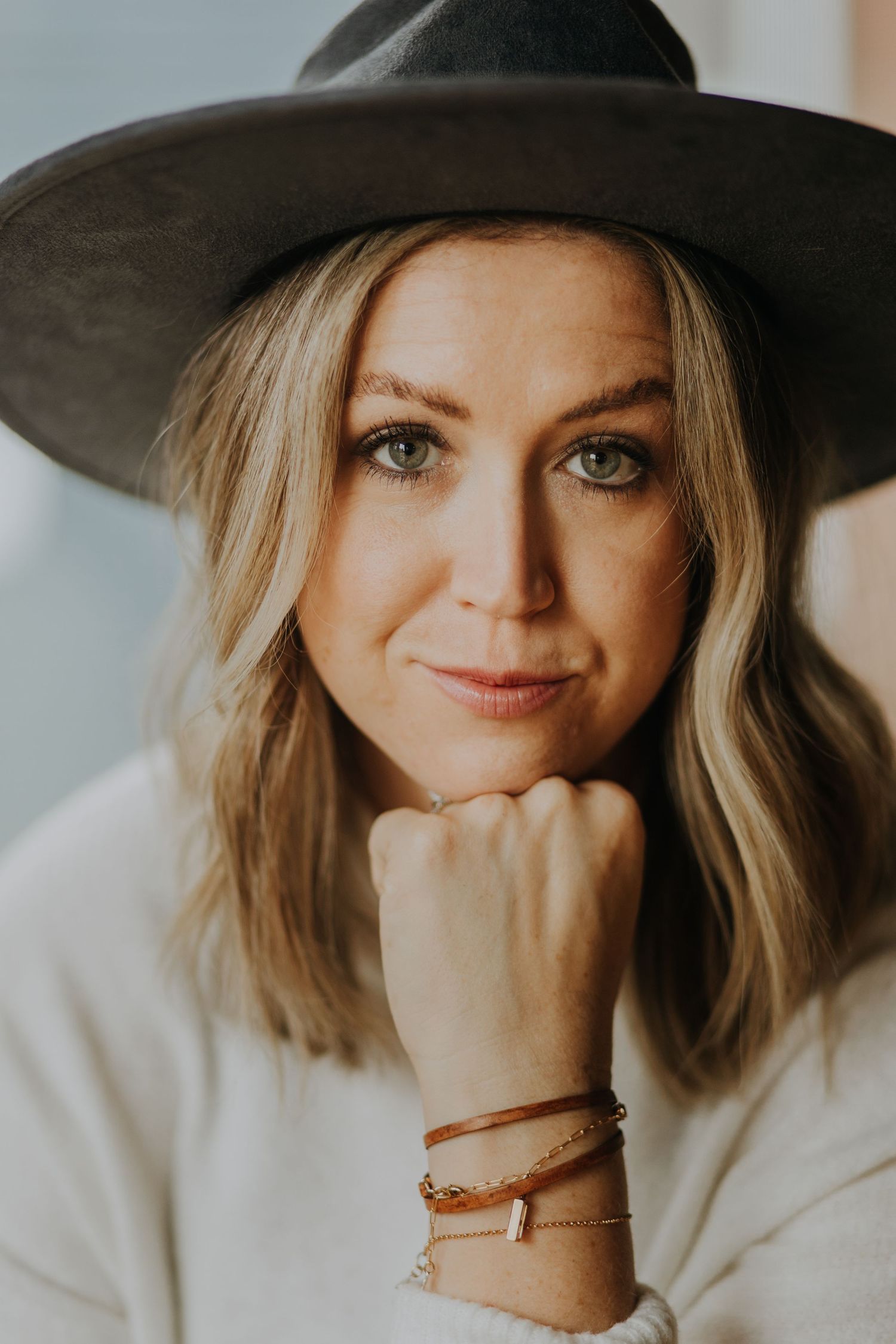 Blonde woman wearing a dark wide-brim hat and layered bracelets posing for a portrait.