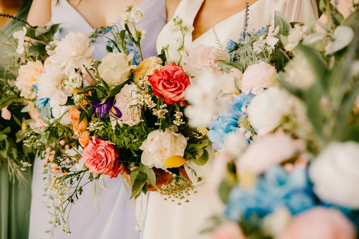 Bridesmaids holding lush colorful wedding bouquets featuring coral roses, white peonies, and blue hydrangeas.