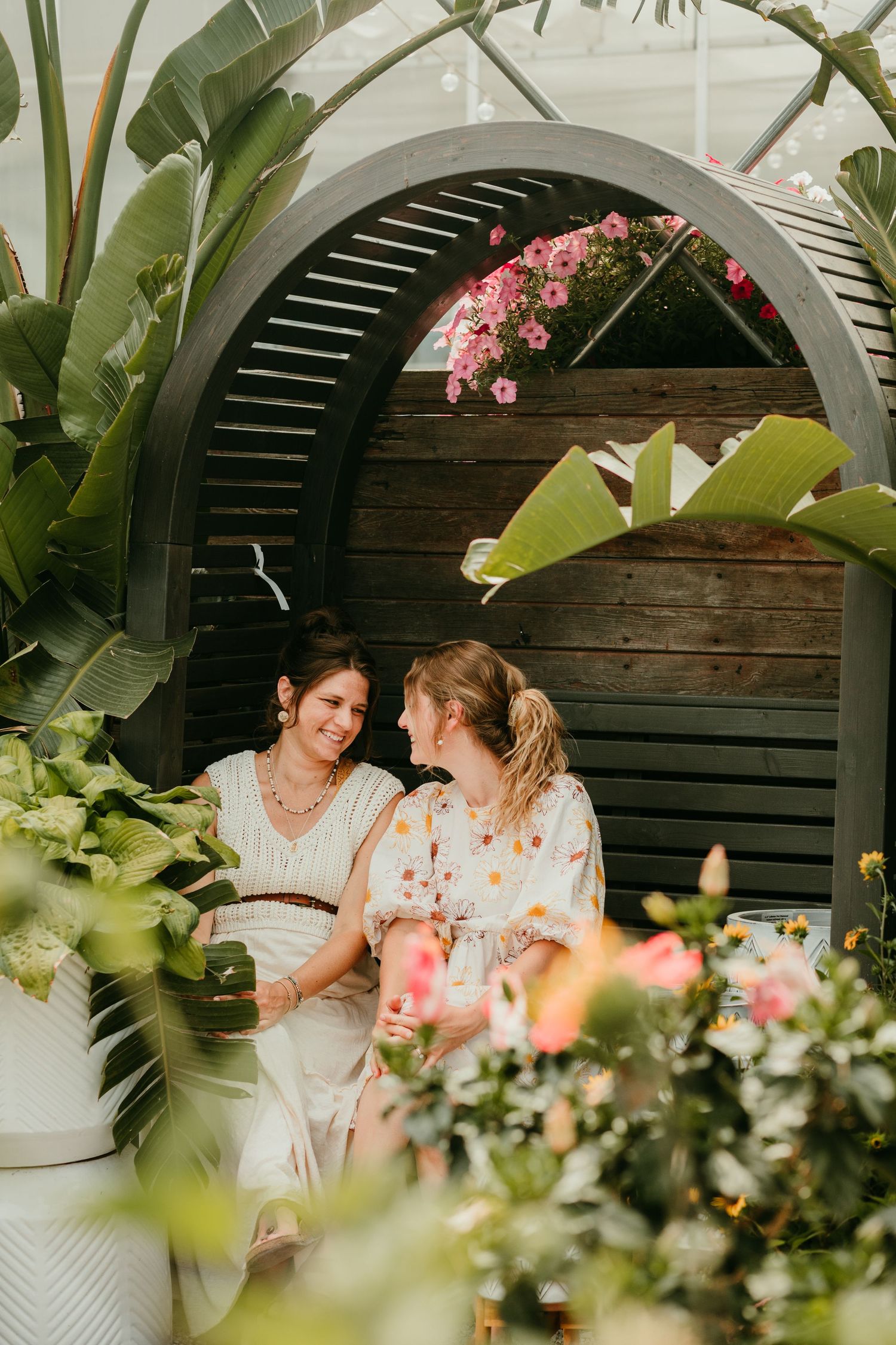 Two women smiling at each other surrounded by lush tropical plants under a wooden arch in a greenhouse.