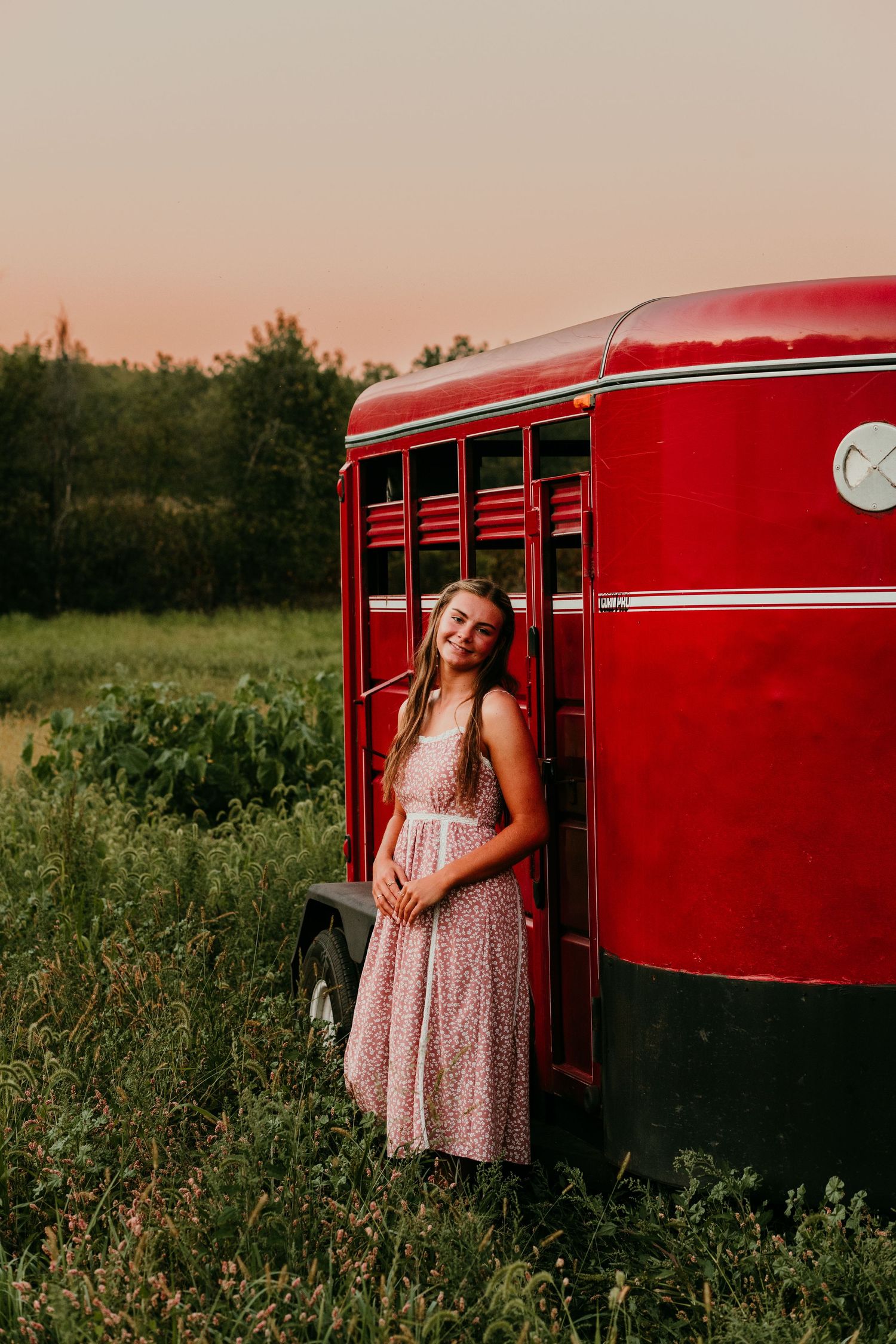 Smiling young woman in floral dress leaning against a vintage red horse trailer at sunset.