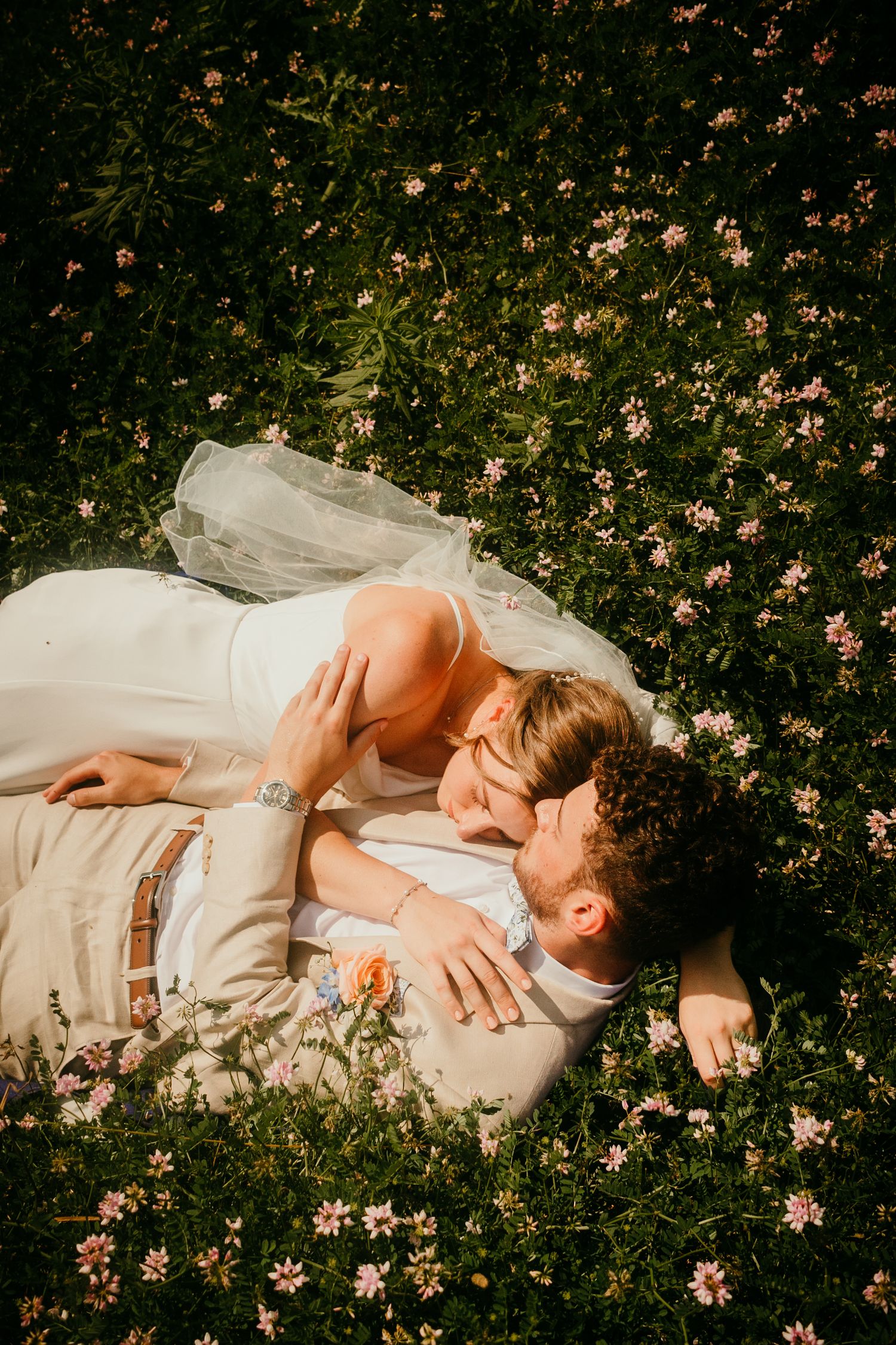 Bride and groom lying in a wildflower meadow sharing a tender kiss during golden hour.