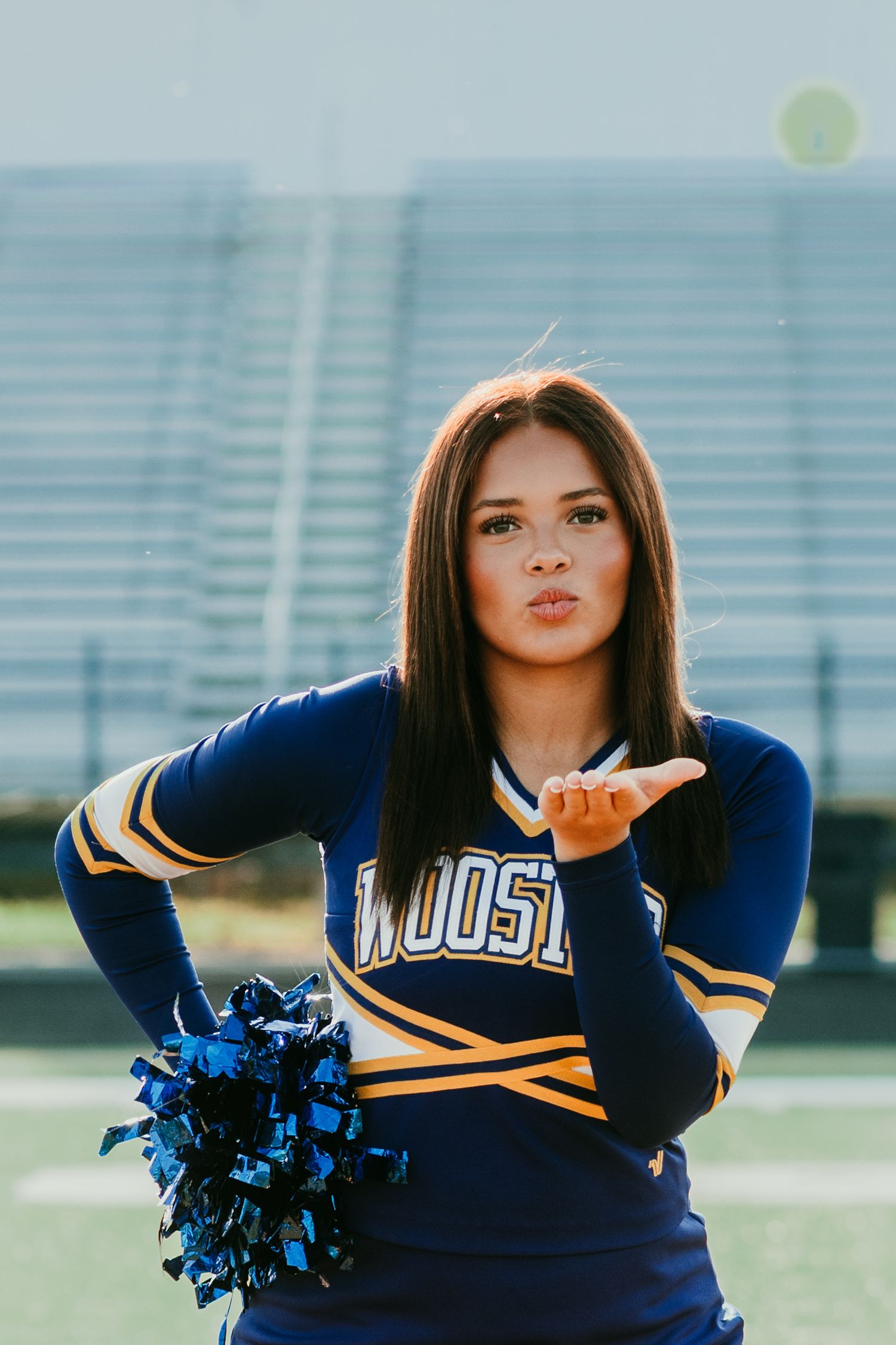 Cheerleader in navy and gold uniform blowing a kiss while holding blue pom-poms on a football field.