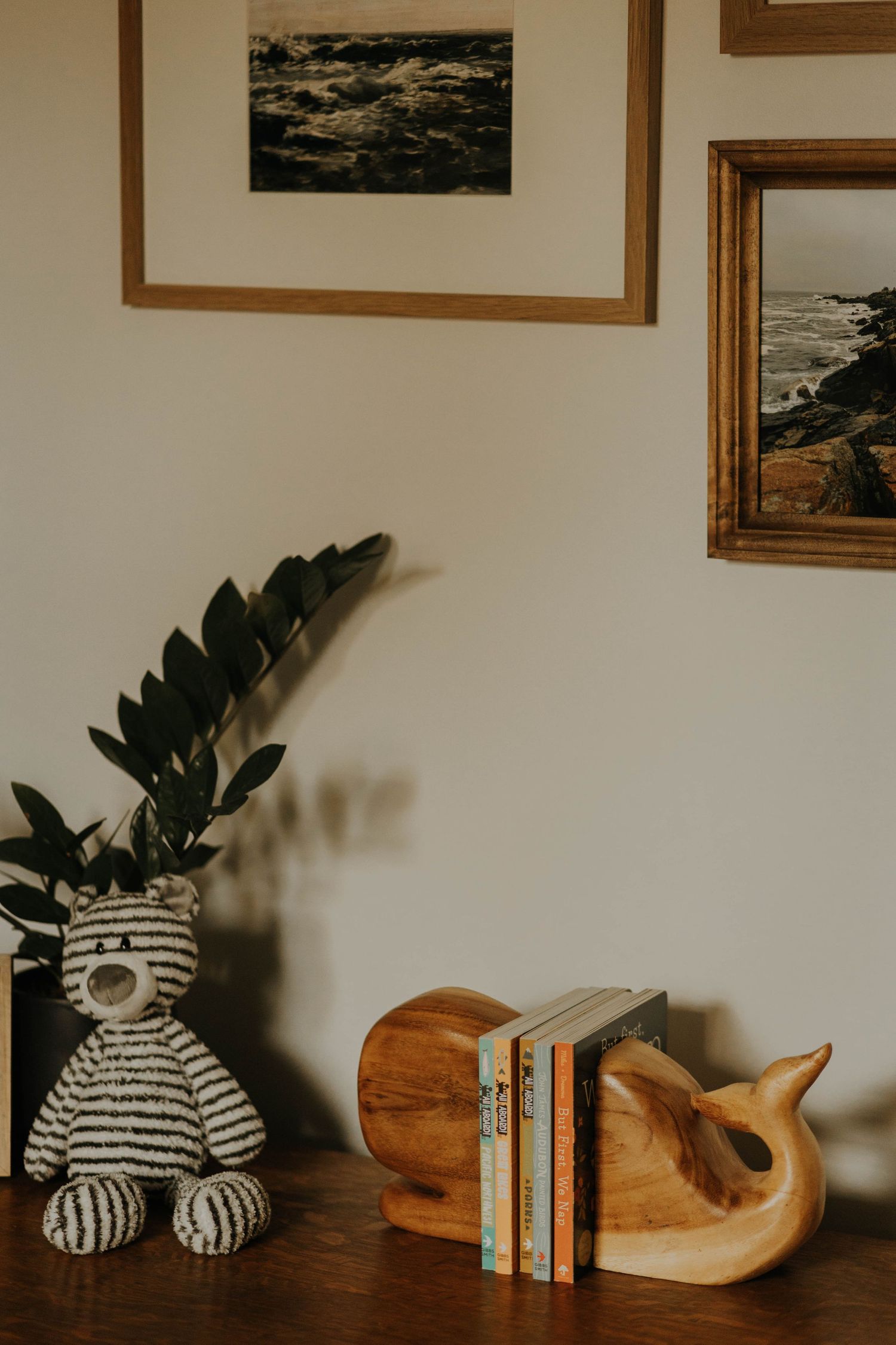 Cozy nursery shelf with striped teddy bear, wooden whale bookends holding childrens books, and framed ocean prints on wall.