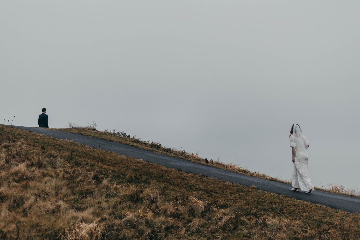 Bride and groom standing apart on a misty hillside road in dramatic landscape.