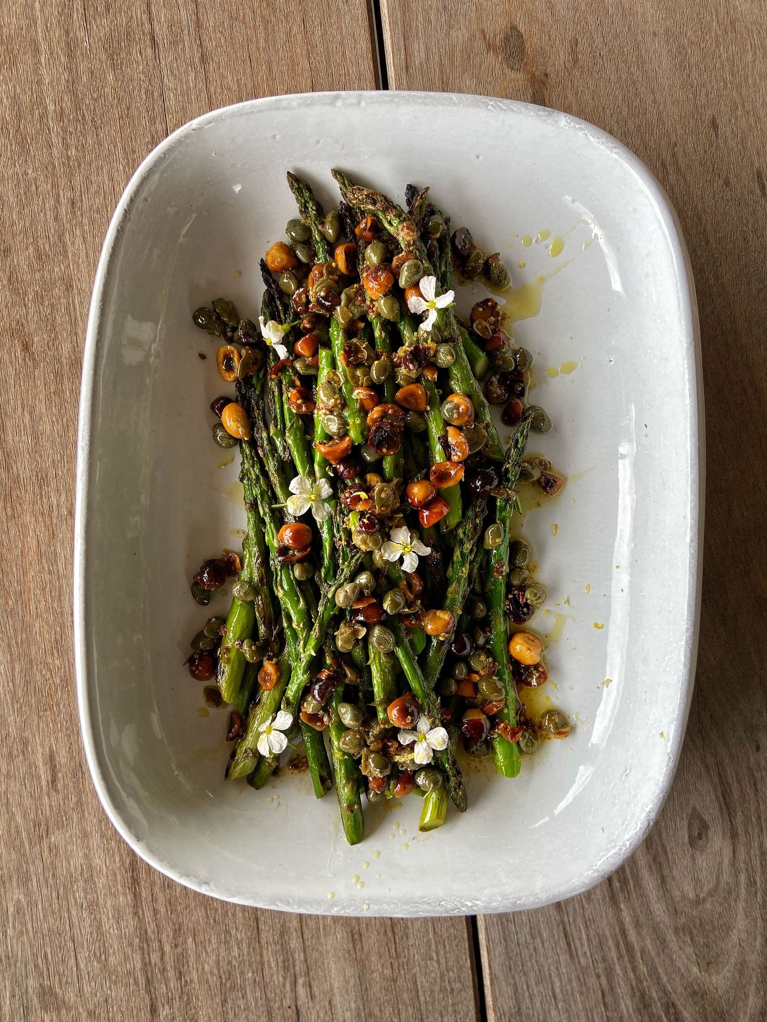Grilled asparagus with crispy capers, dried tomatoes, and white flowers in a white ceramic dish.