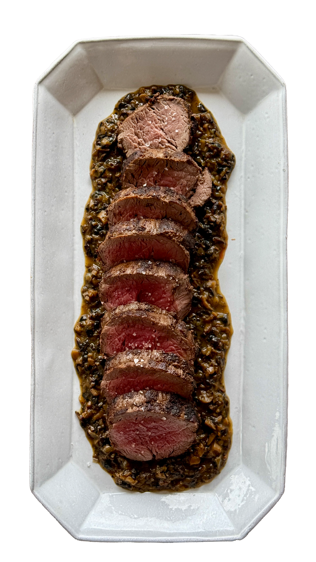 Sliced medium-rare beef tenderloin on mushroom sauce served on white rectangular platter.