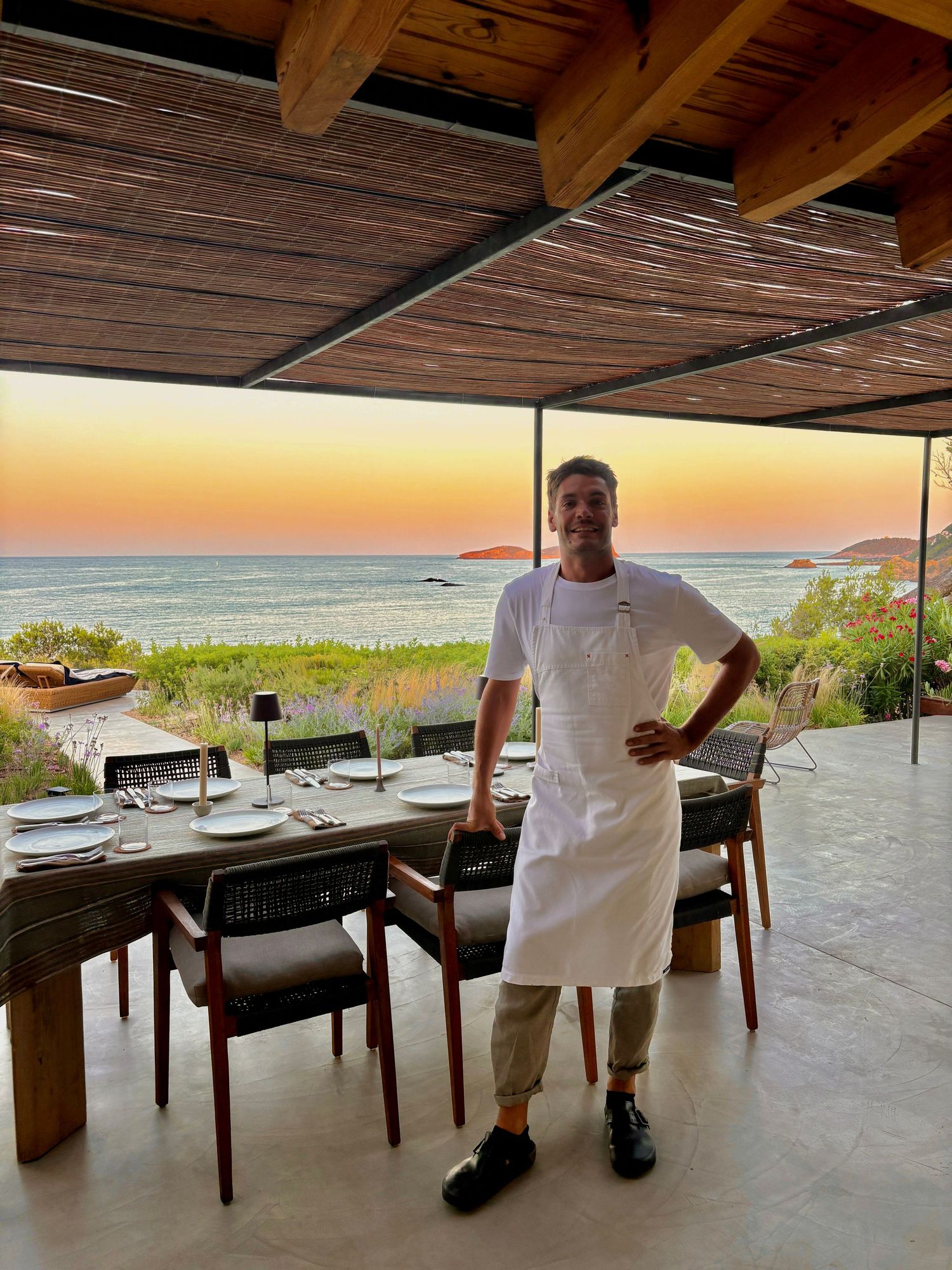 Chef in white apron standing by a set dining table with stunning ocean sunset view.