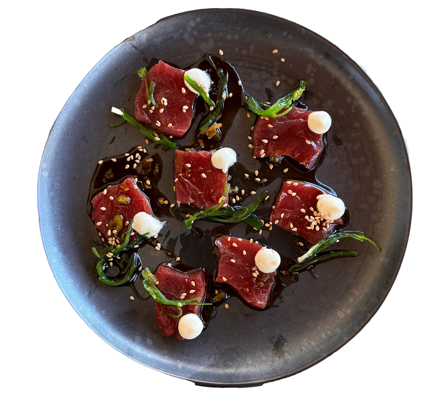 Fresh tuna sashimi with sesame seeds, scallions, white cream dots, and dark sauce on a dark ceramic plate.