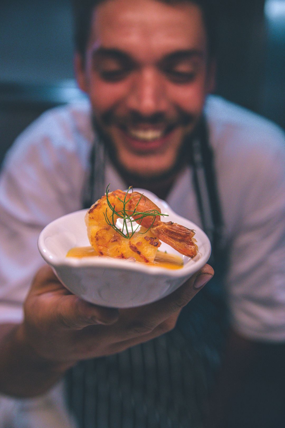 Smiling chef presenting a perfectly cooked whole shrimp with dill garnish in a small white ceramic bowl.