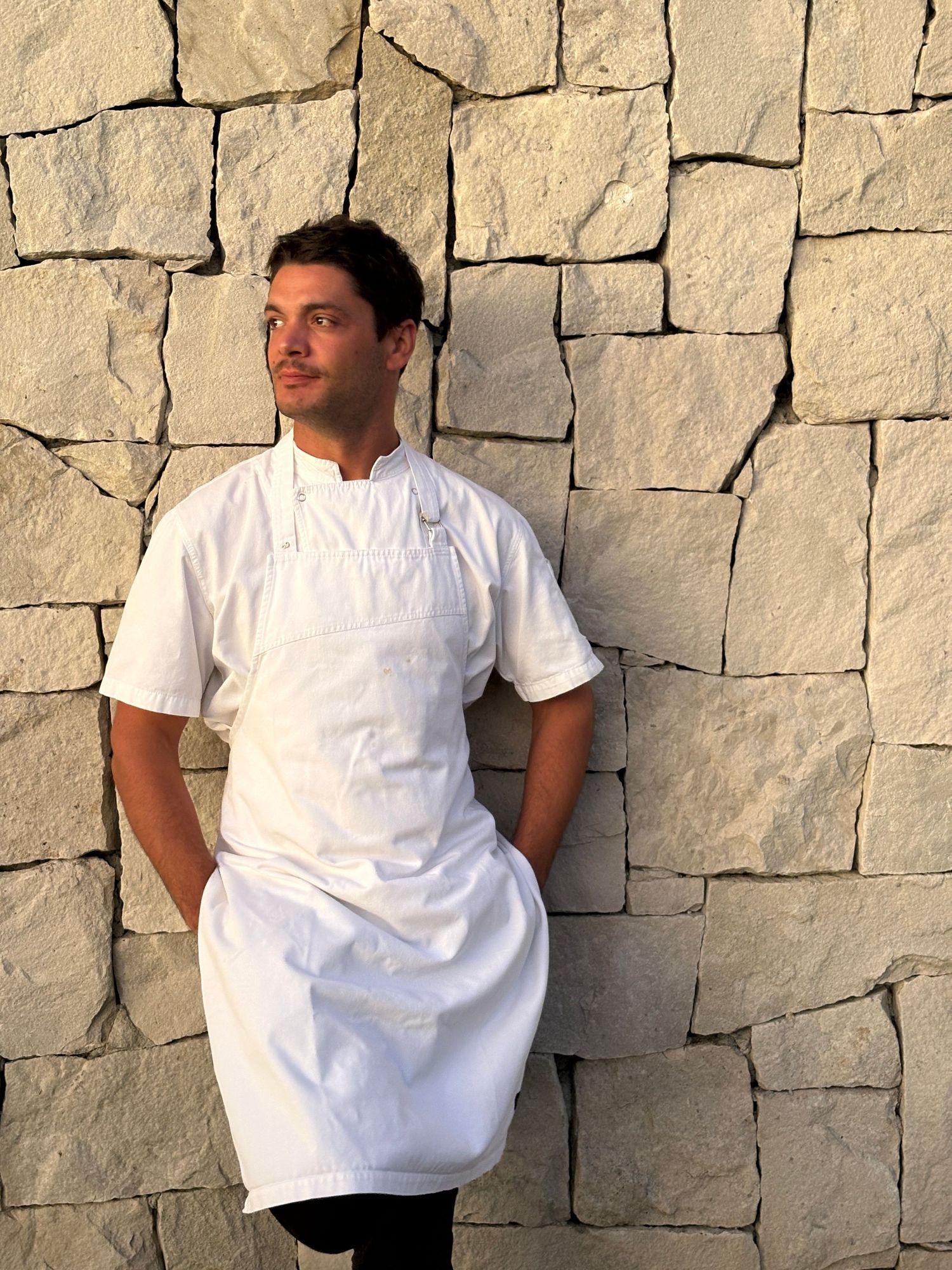 Young male chef in white uniform and apron leaning against a stone wall outdoors.