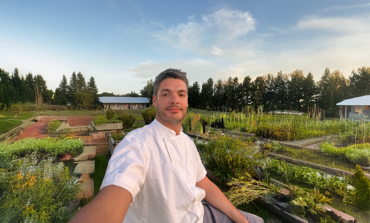 Private chef in white uniform taking a selfie in a lush garden with trees in background.