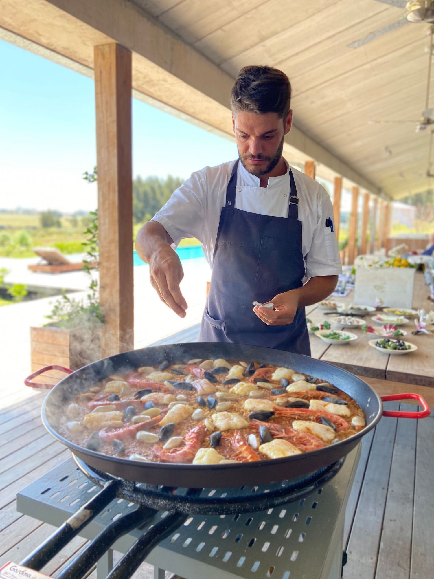 Chef in apron seasoning a large paella pan filled with seafood on an outdoor terrace.