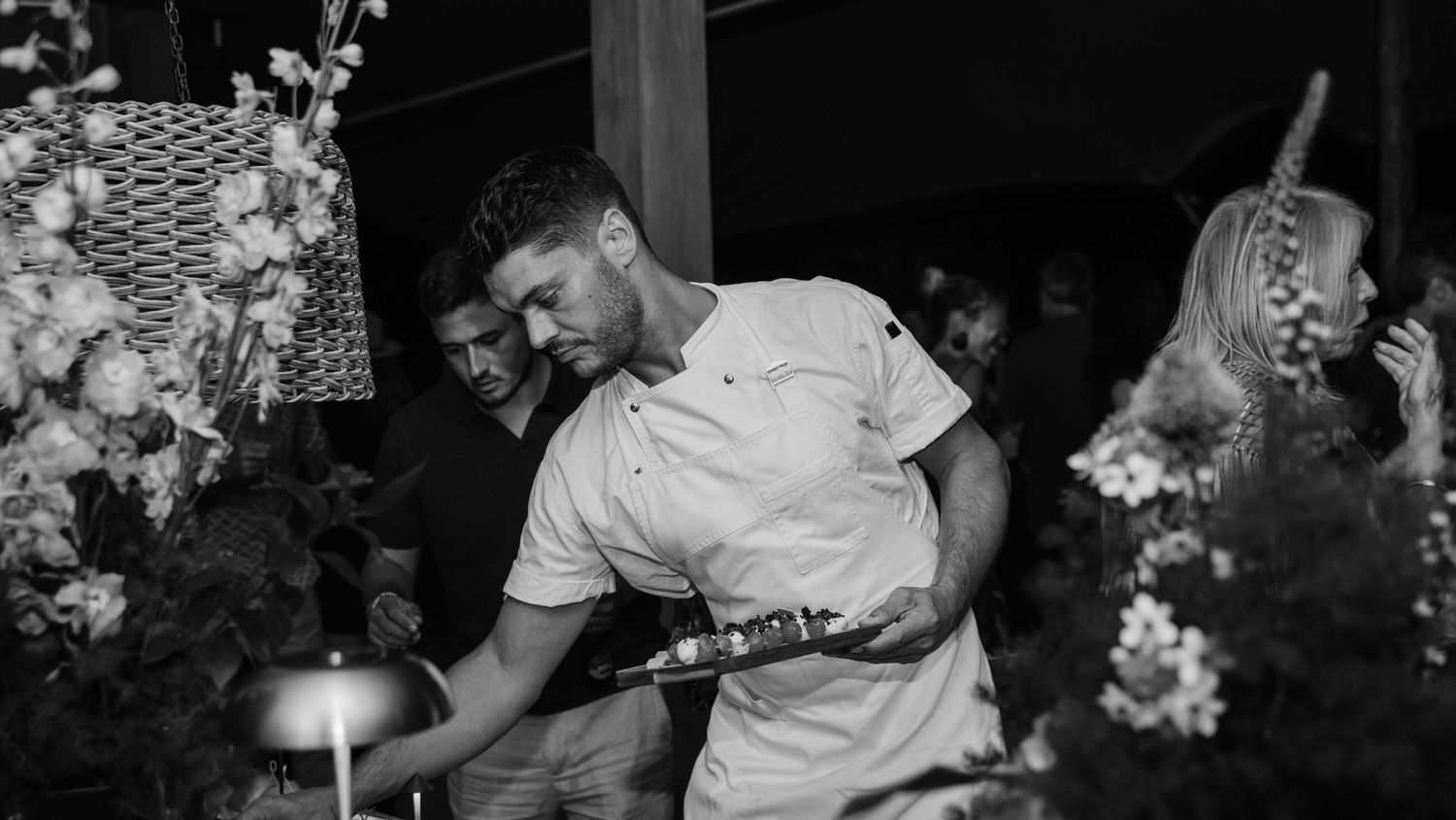 Chef in white uniform pouring a drink at an elegant outdoor event surrounded by floral arrangements.