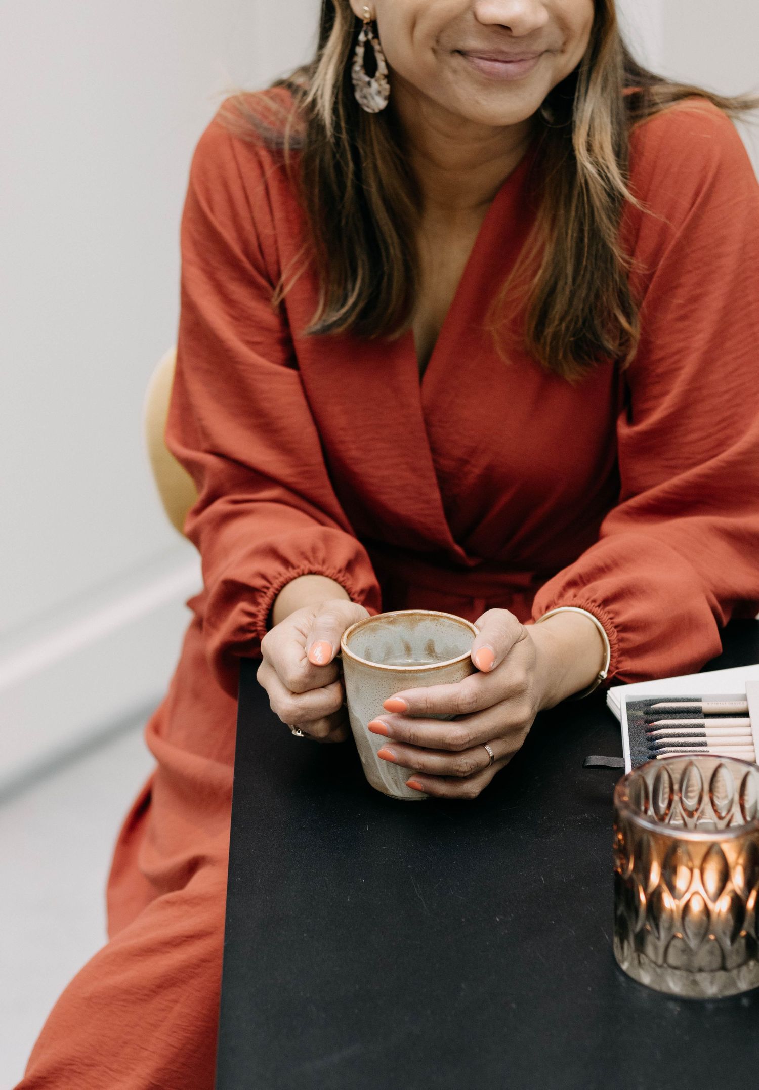 Woman in red wrap dress holding a ceramic mug at a dark table with a candle nearby.
