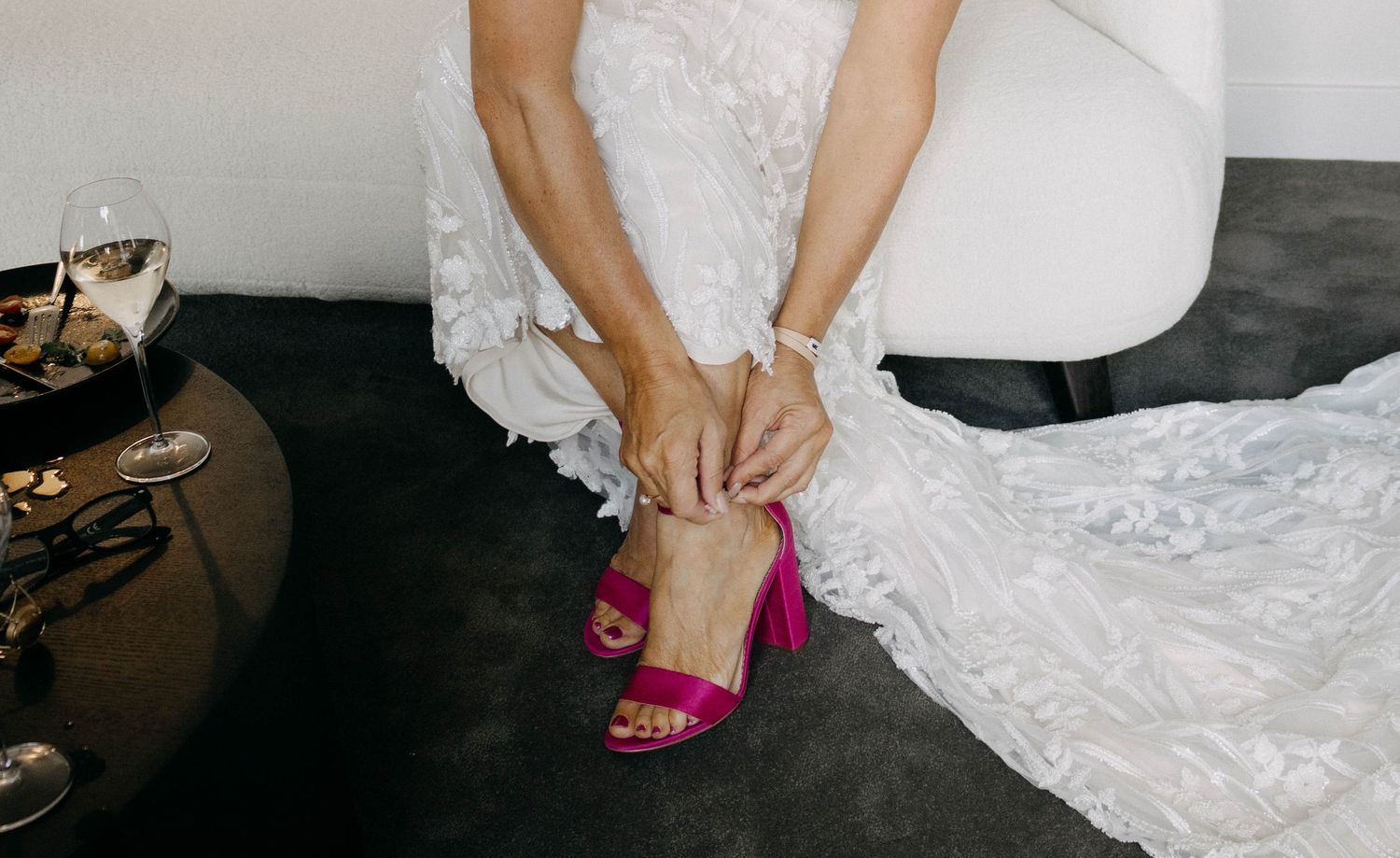 Bride in white lace wedding dress adjusting pink block heel sandals while seated on a chair.