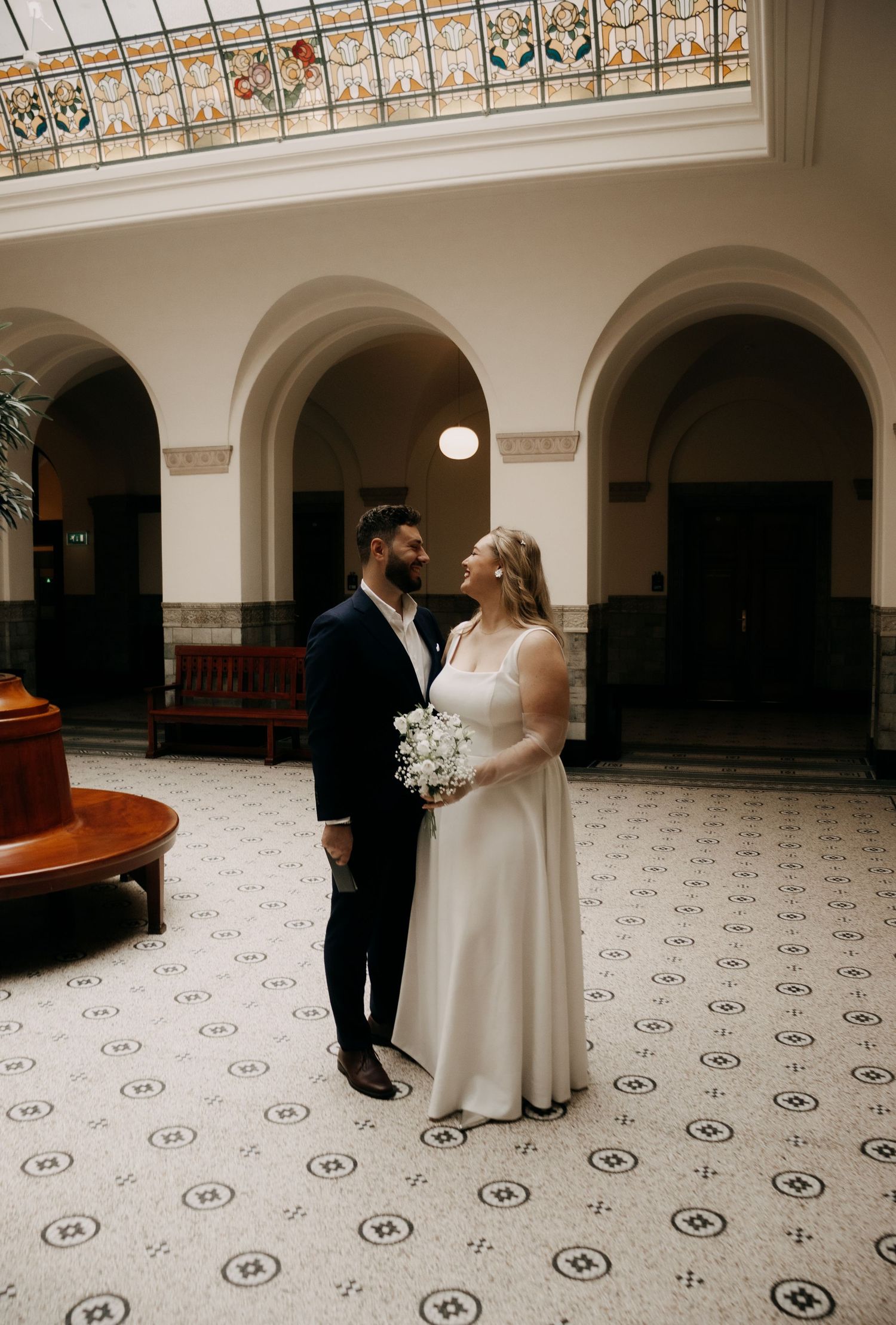 Bride and groom share a romantic moment in an elegant hall with ornate tiled floors and stained glass ceiling.