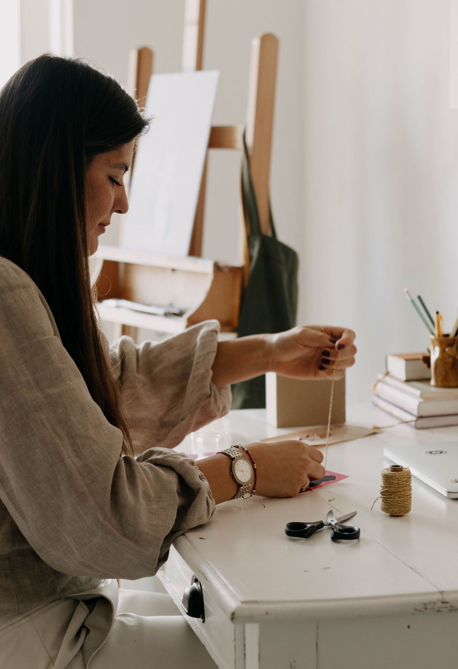 Woman crafting at a white desk with twine, art supplies, and an easel in a bright studio.
