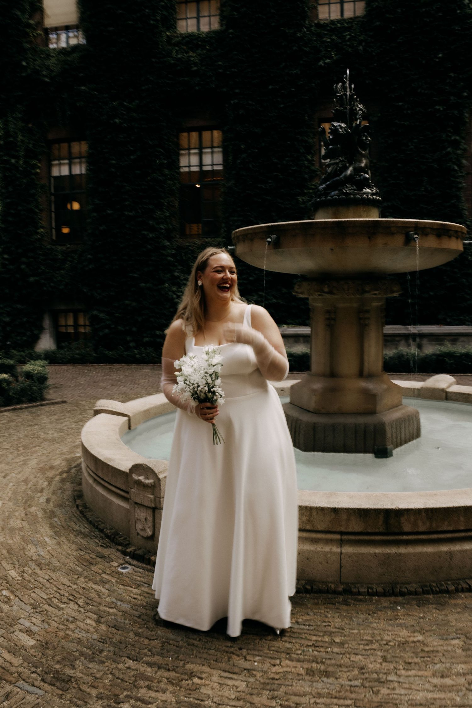 Joyful bride in white gown laughing while holding bouquet beside ornate stone fountain outdoors.