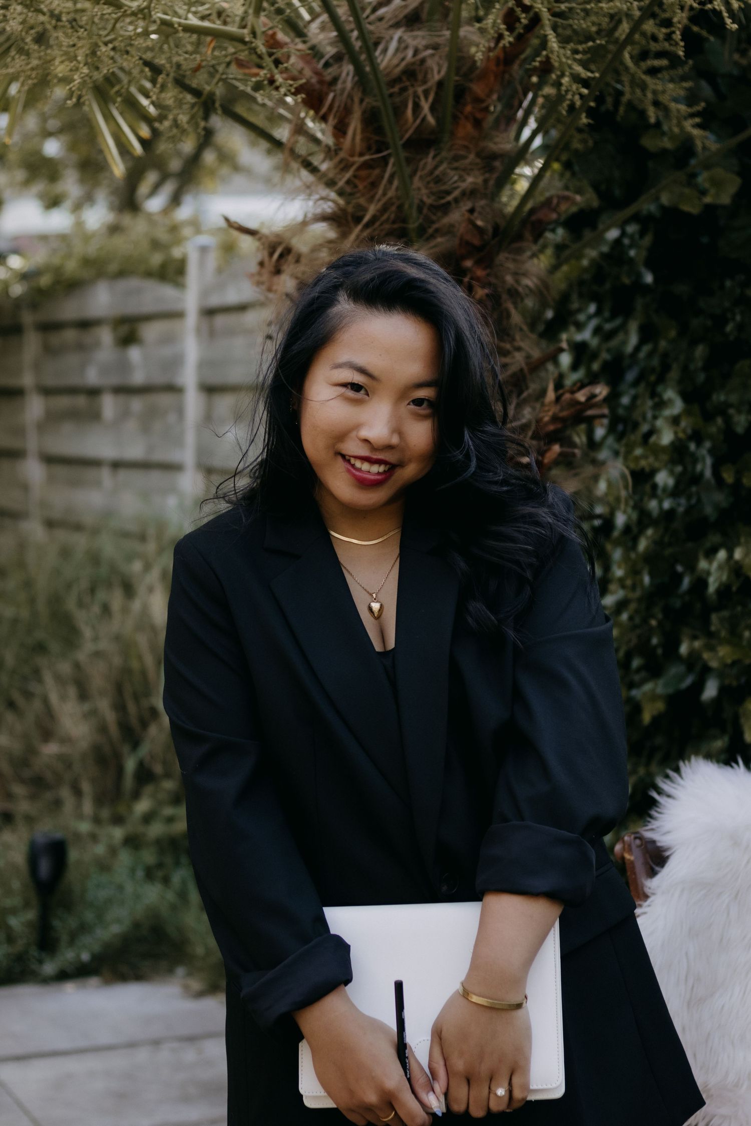 Smiling Asian woman in black blazer holding a white folder outdoors among green foliage.