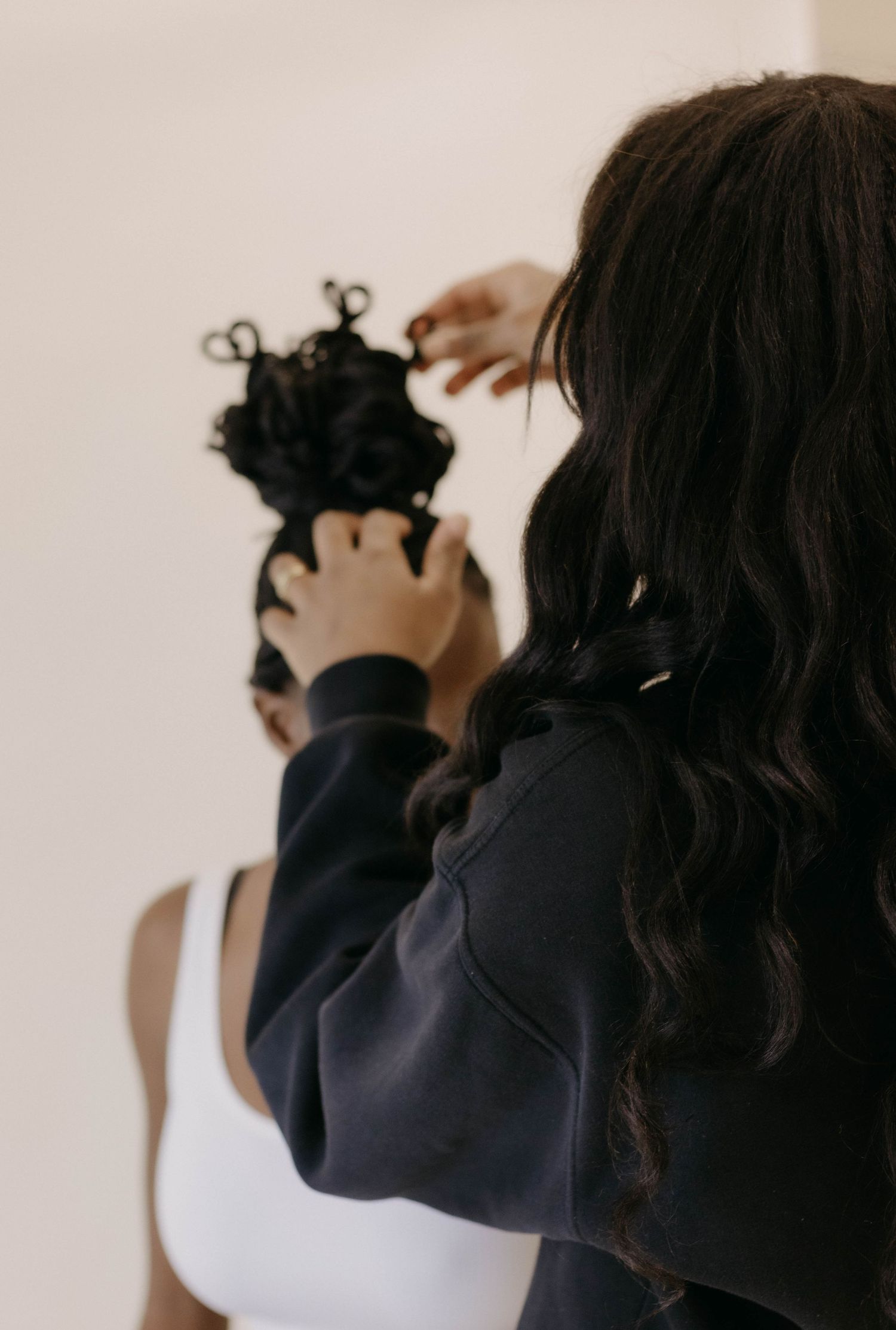 A hairstylist with long wavy hair styling another woman's curly updo from behind.