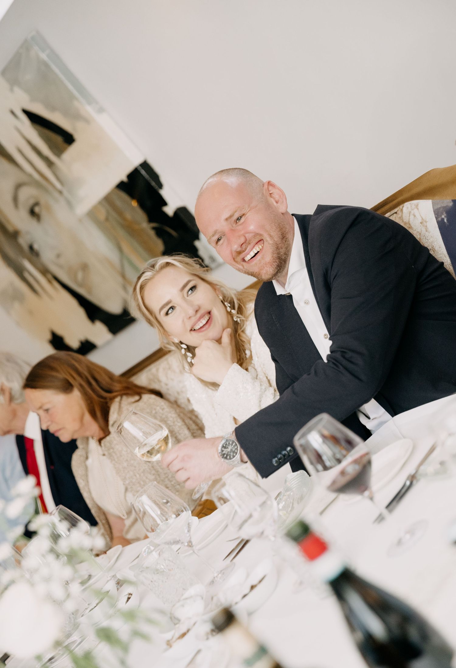 Smiling bride and groom posing together at a wedding reception dinner table with wine glasses and candles.