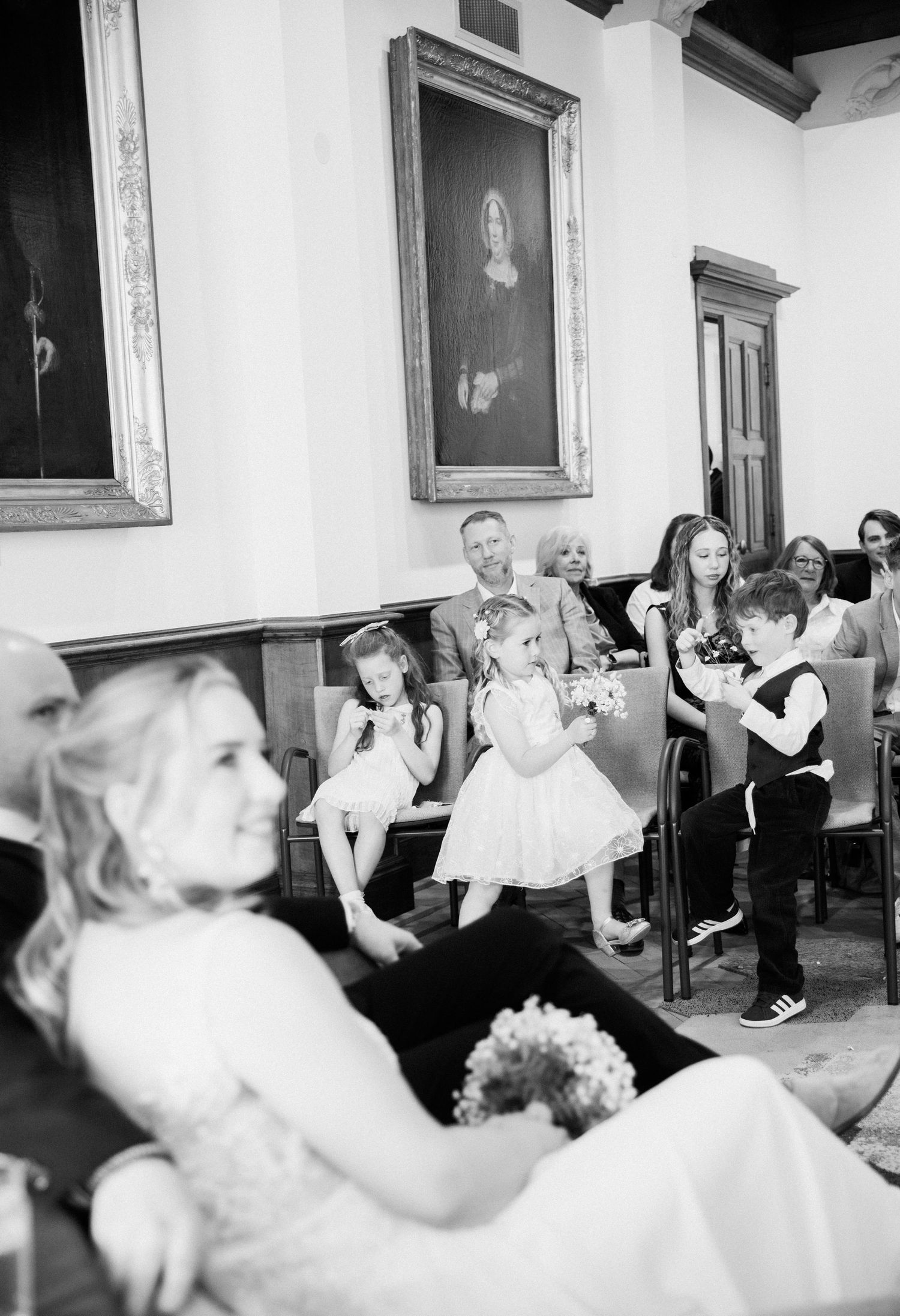 Black and white photo of flower girls and ring bearer seated at wedding ceremony in historic venue.