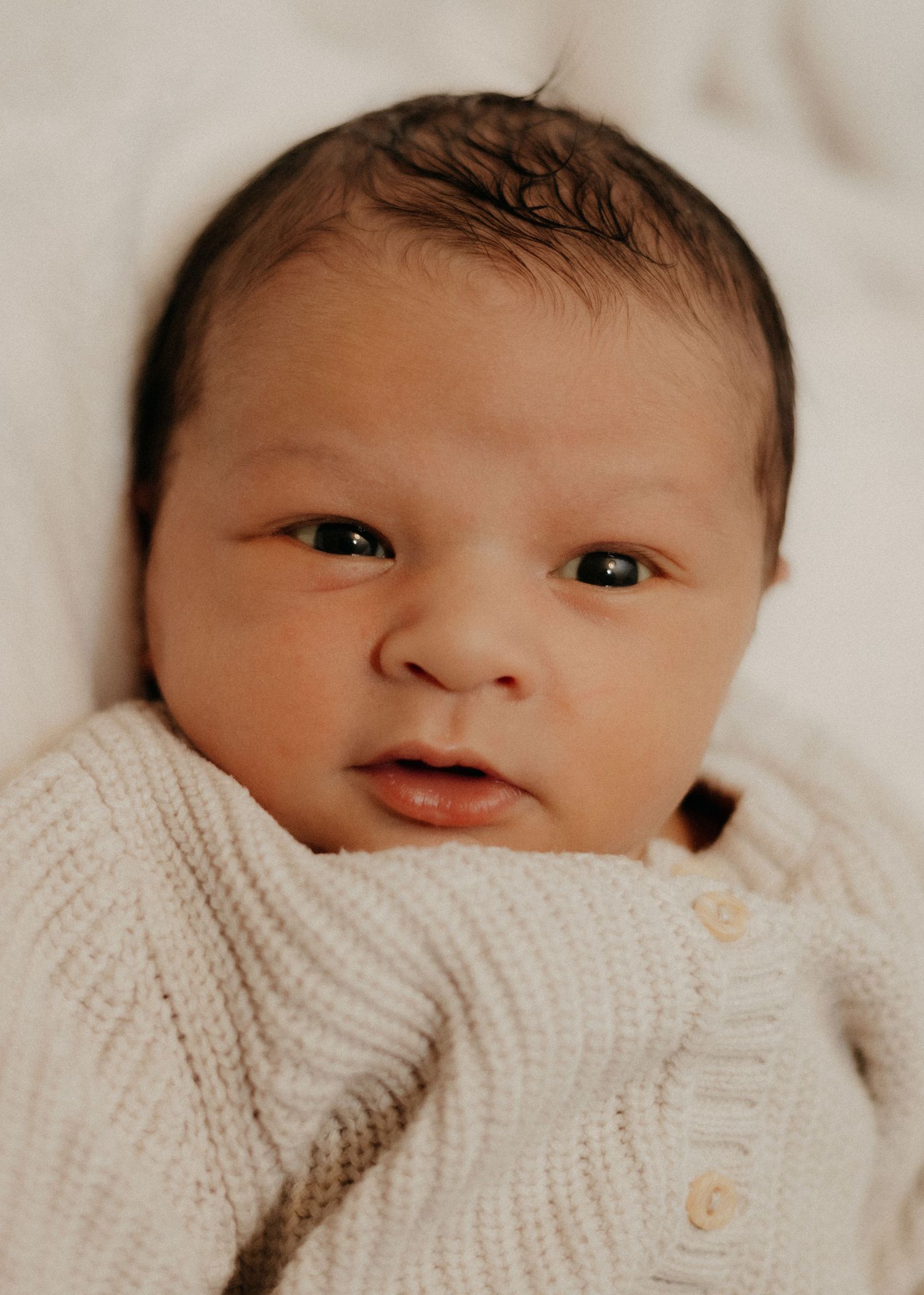 Close-up portrait of a newborn baby with dark hair wrapped in a cream knit blanket.