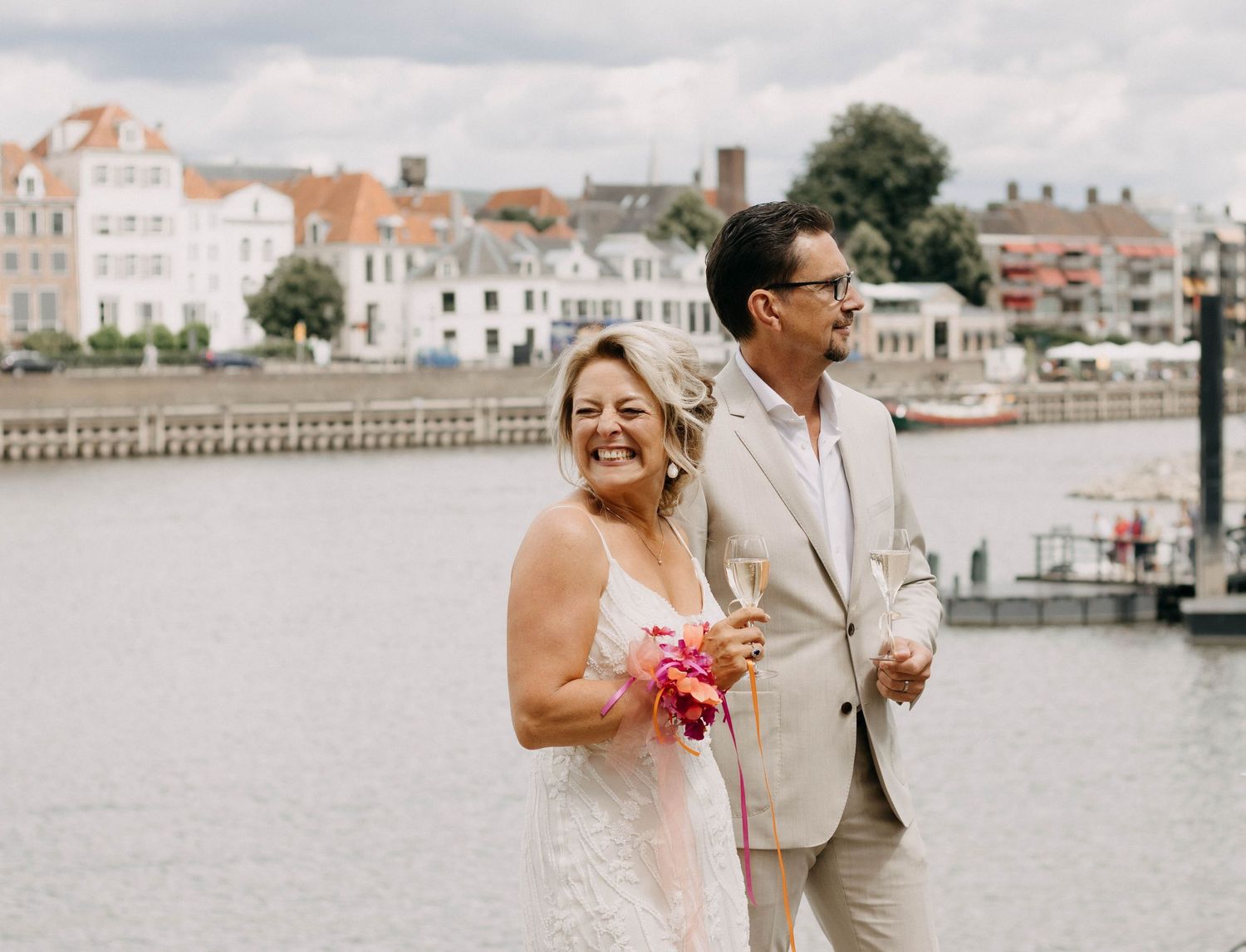 Bride and groom smiling by a waterfront harbor, she holds pink flowers in a white gown, he wears a beige suit.
