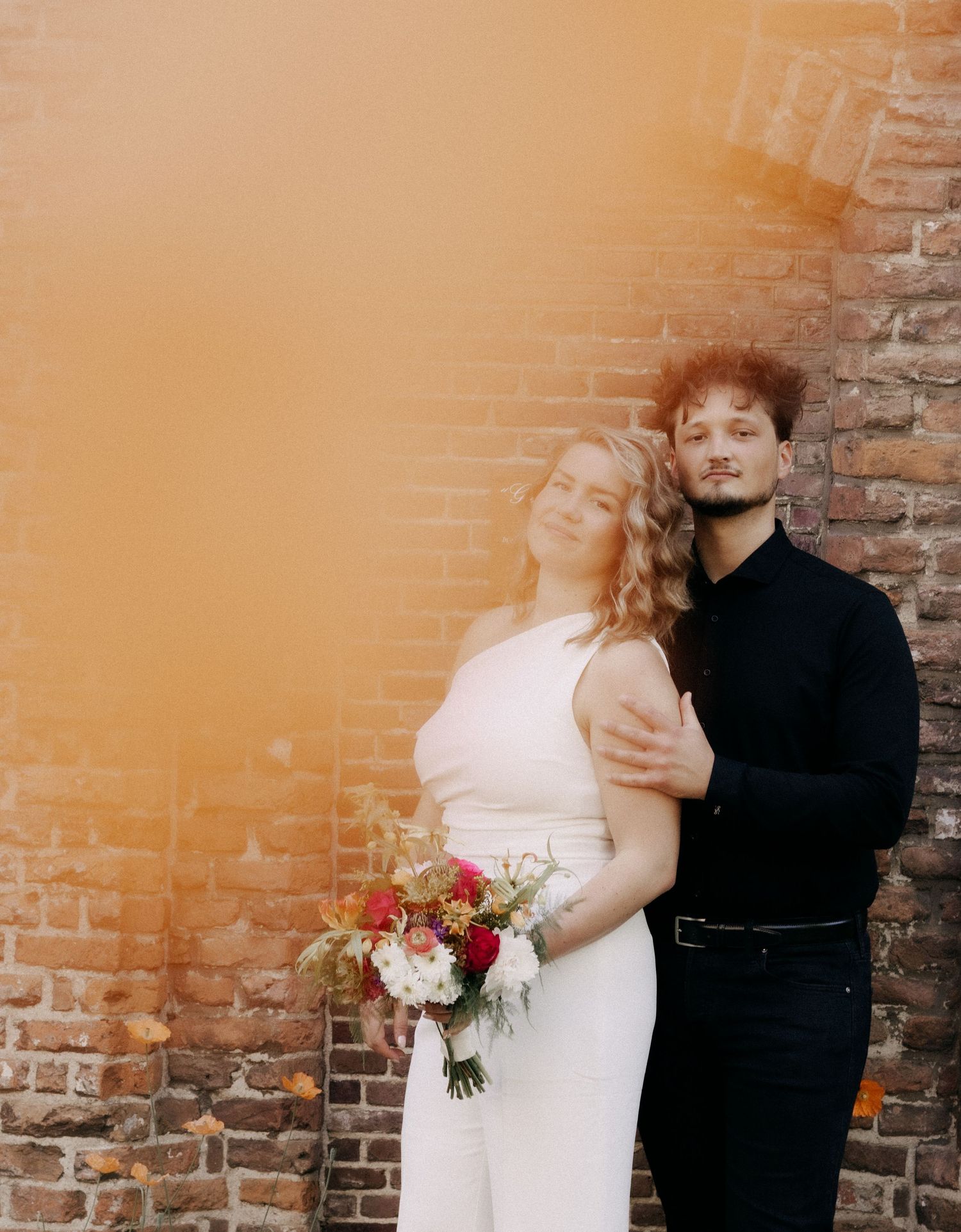 Couple posing against brick wall with orange smoke, bride in white holding colorful bouquet.