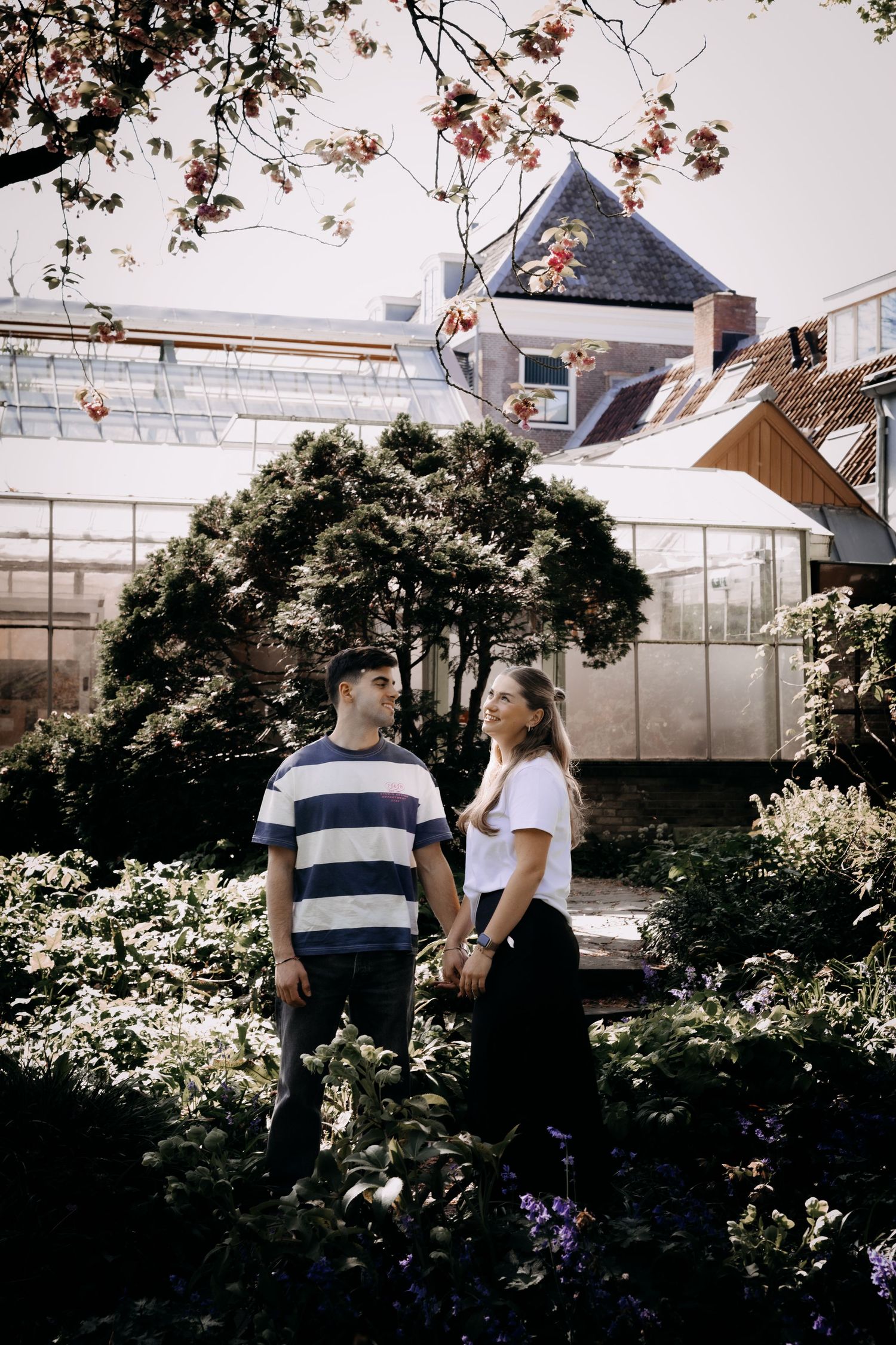 Couple holding hands and smiling at each other in a lush garden with greenhouse backdrop.
