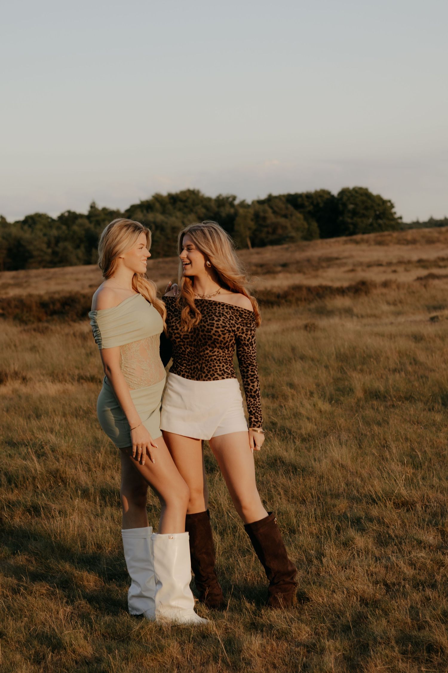 Two blonde women in stylish outfits posing in a sunlit meadow wearing boots.