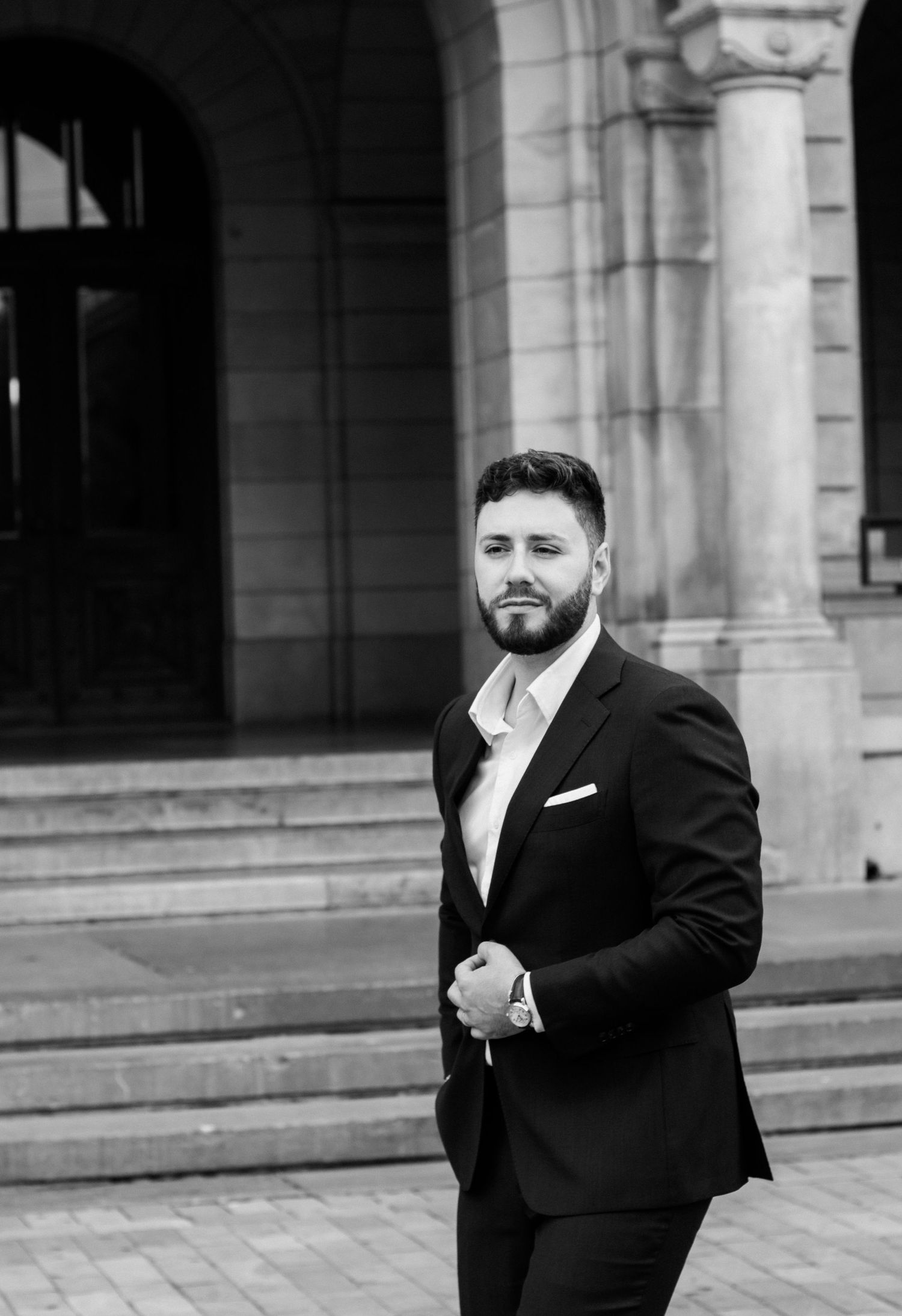 Dapper groom in black suit with pocket square posing confidently outside a stone building.